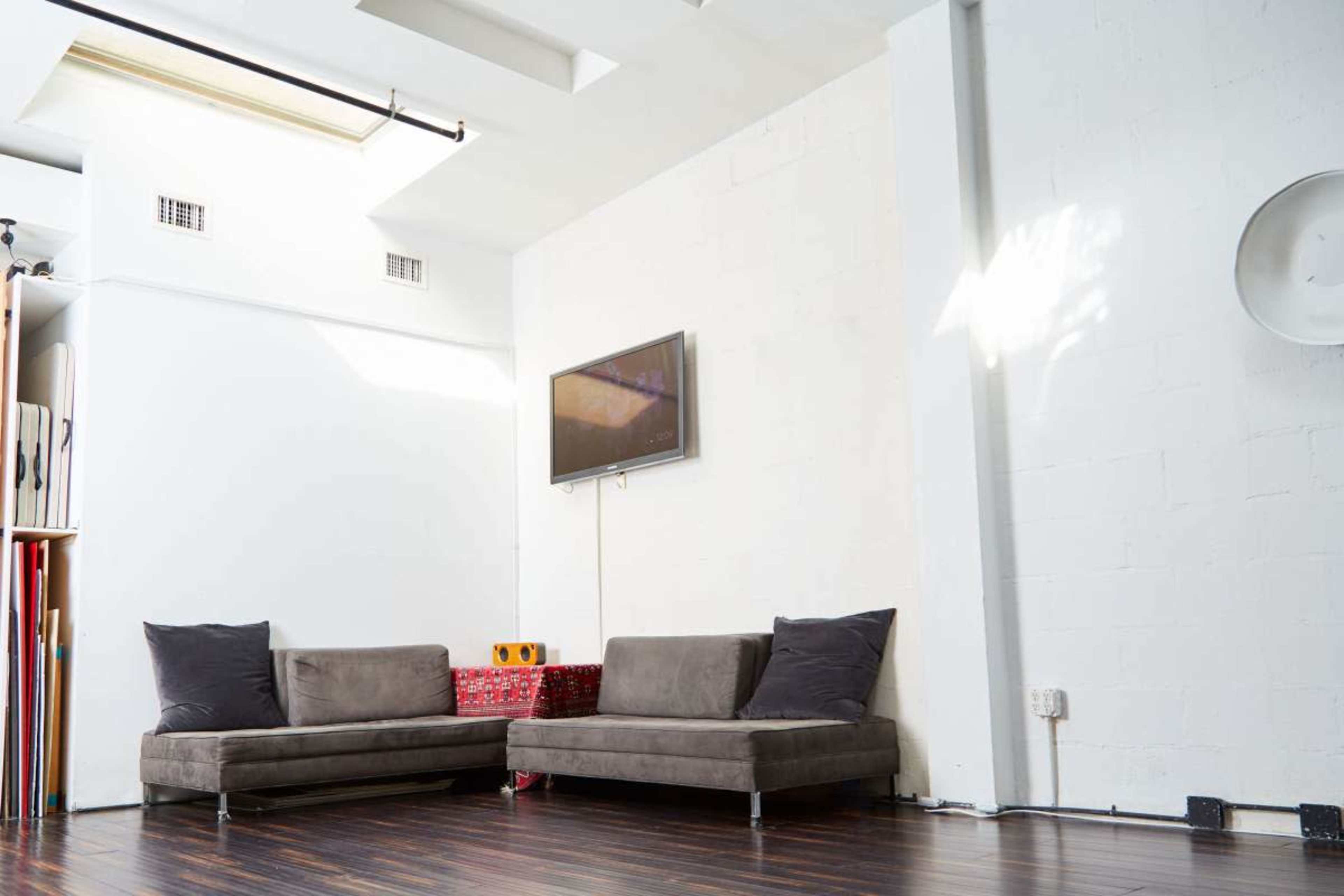 The image shows a minimalist interior with two gray sofas situated against a white wall, a television mounted above them, and wooden flooring.