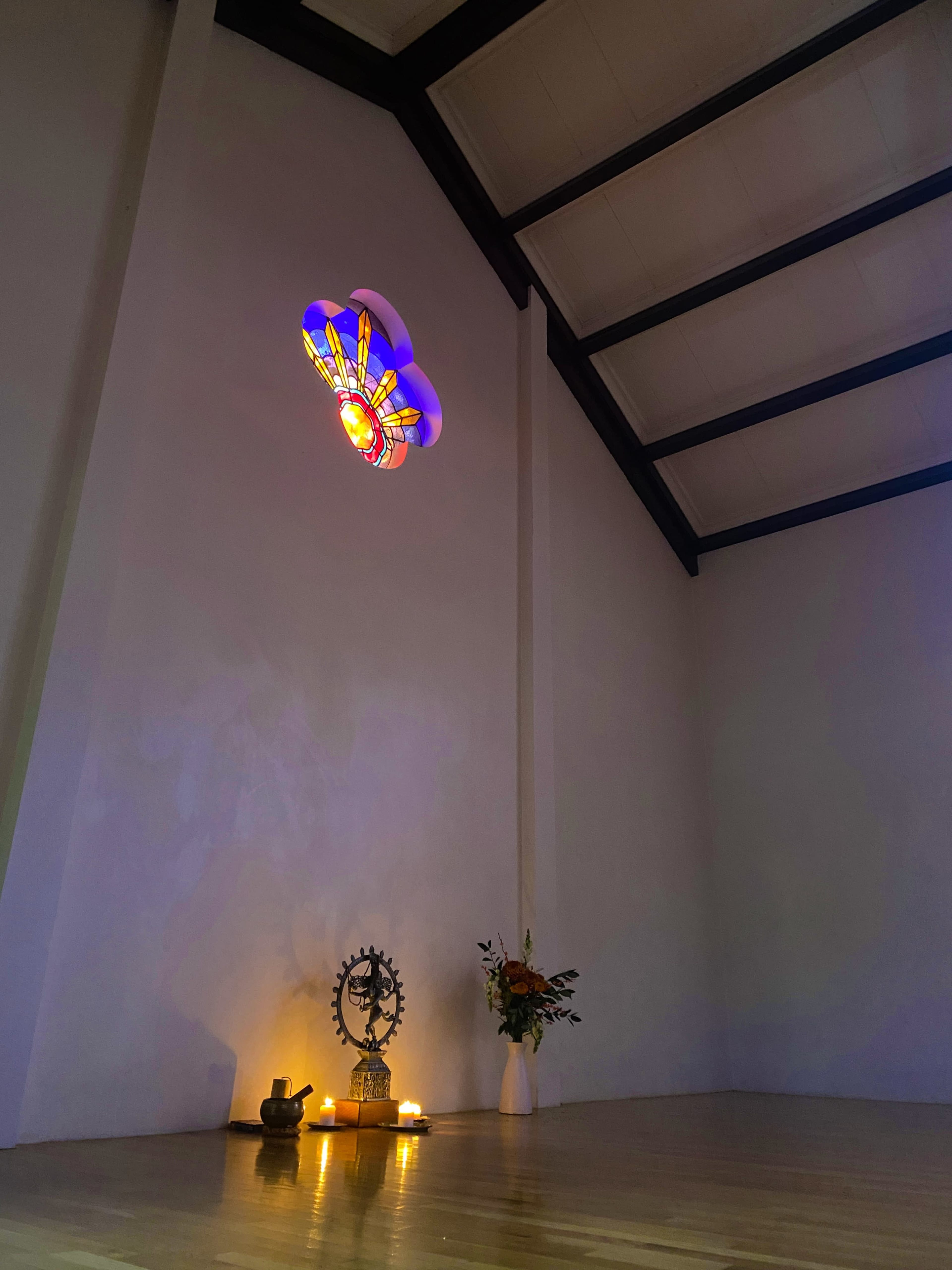 The image shows a serene interior space featuring a stained glass window, a decorative altar with candles, and a flower arrangement.