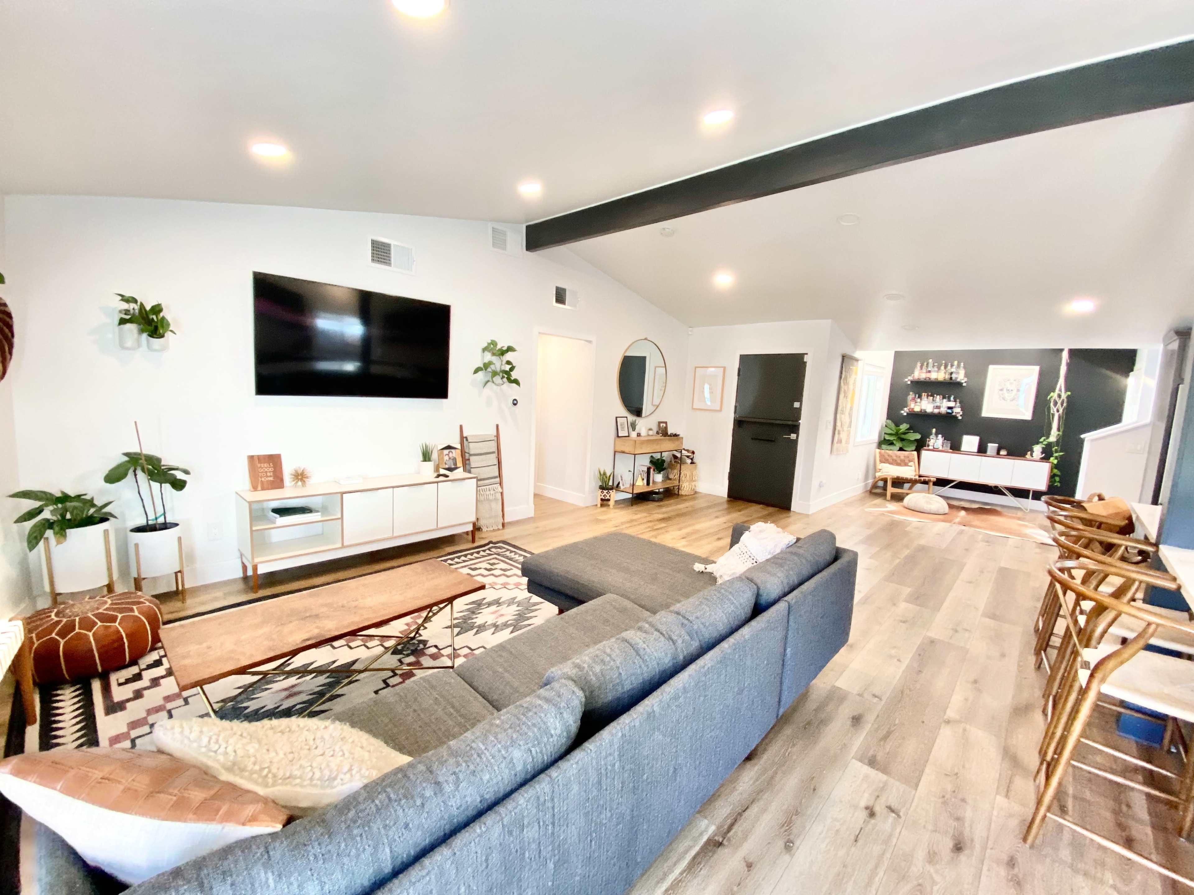 The image shows a modern living room with a sectional couch, a large television on the wall, and a dining area visible in the background.