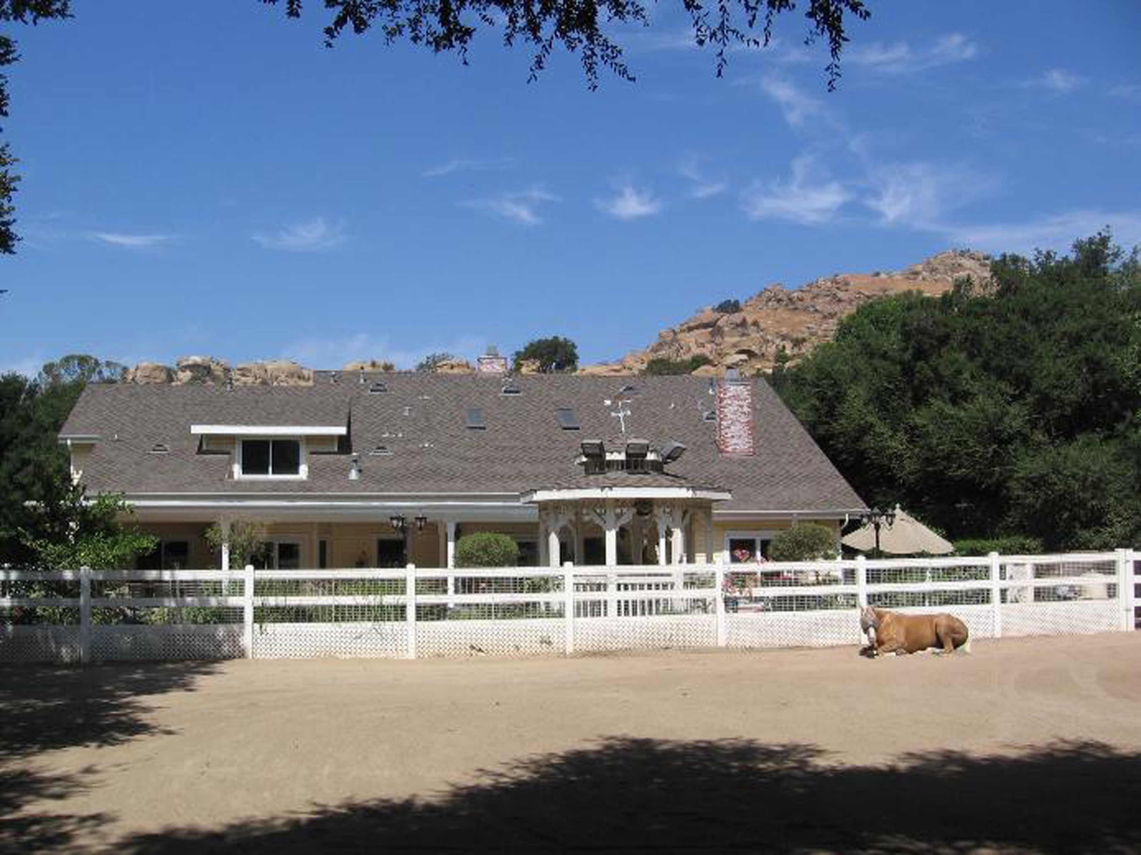 A large house with a brown roof and white fence is set against a rocky hillside under a blue sky.