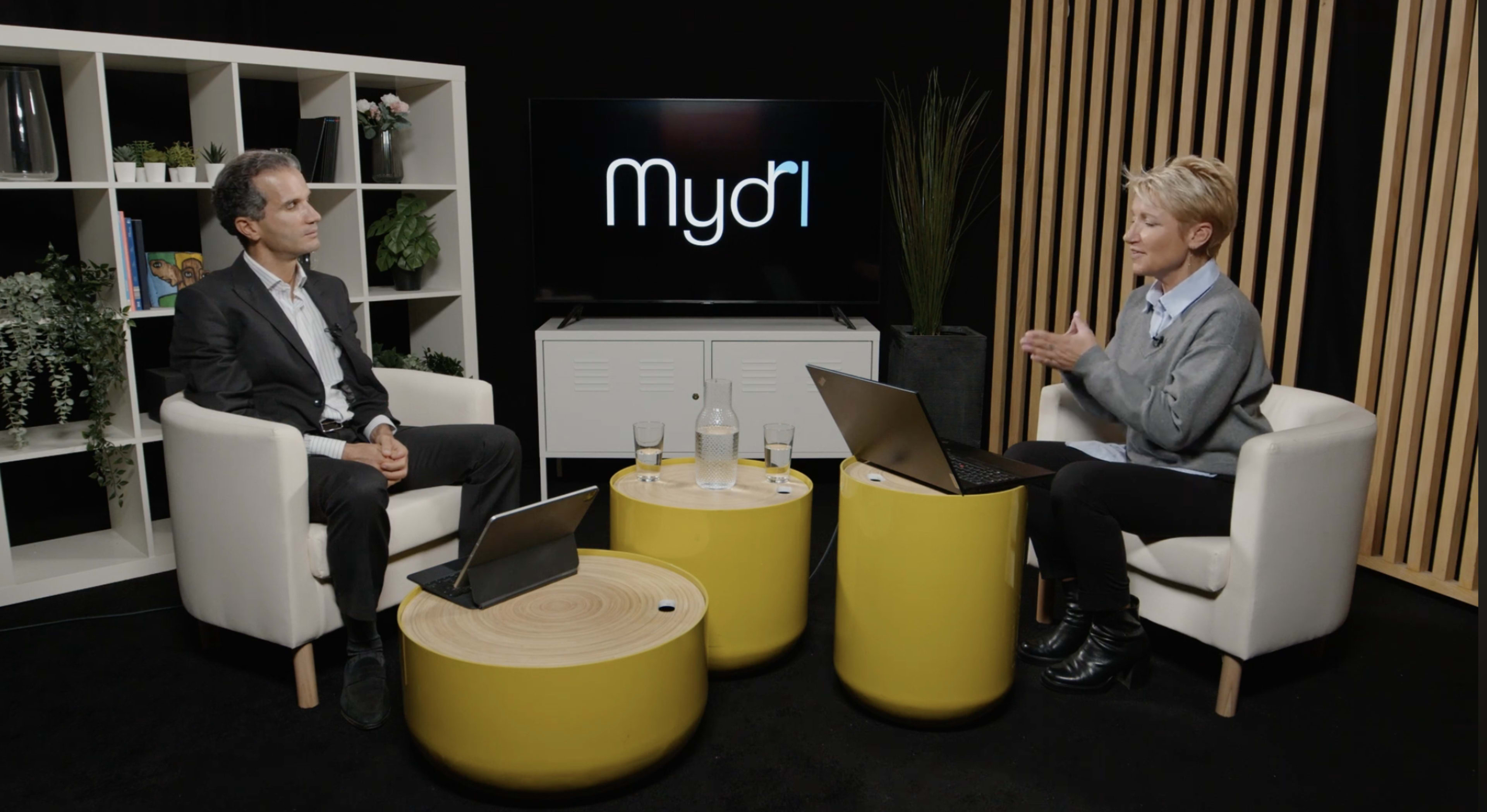 A man and a woman sit in modern chairs facing each other, with a laptop on a yellow coffee table between them, in a studio setting featuring plants and a television screen displaying the logo "Mydri."