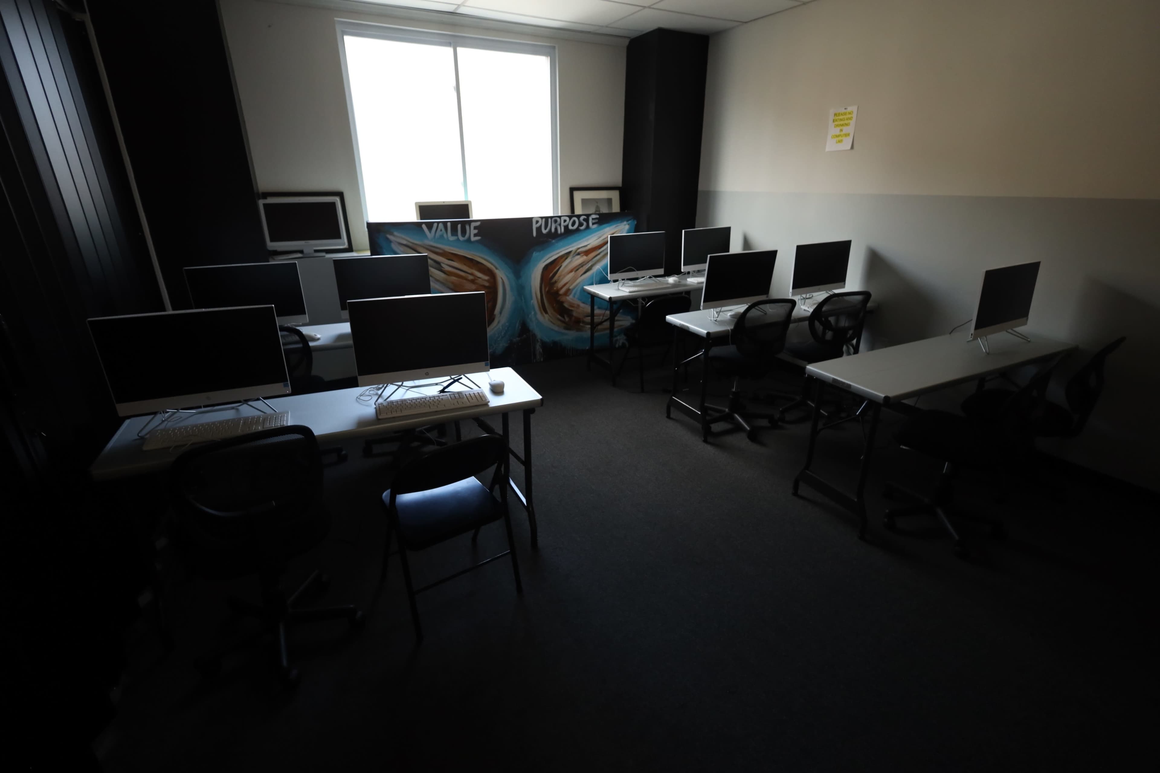 The image shows a small, dimly lit classroom equipped with multiple computer stations arranged around a mural that reads "VALUE PURPOSE."