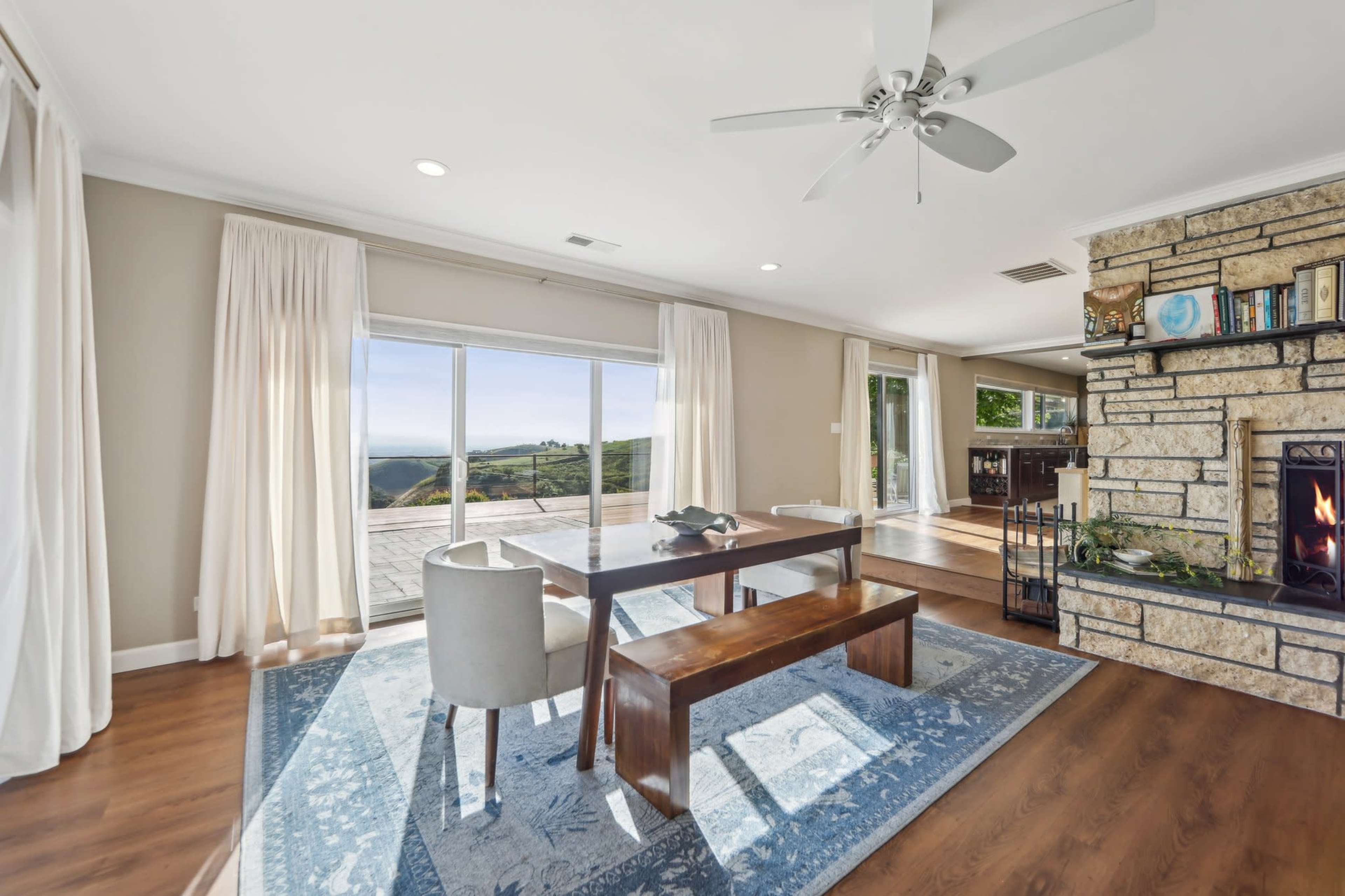 A spacious living area features a wooden dining table with a bench, a stone fireplace, and large windows that overlook a scenic landscape.