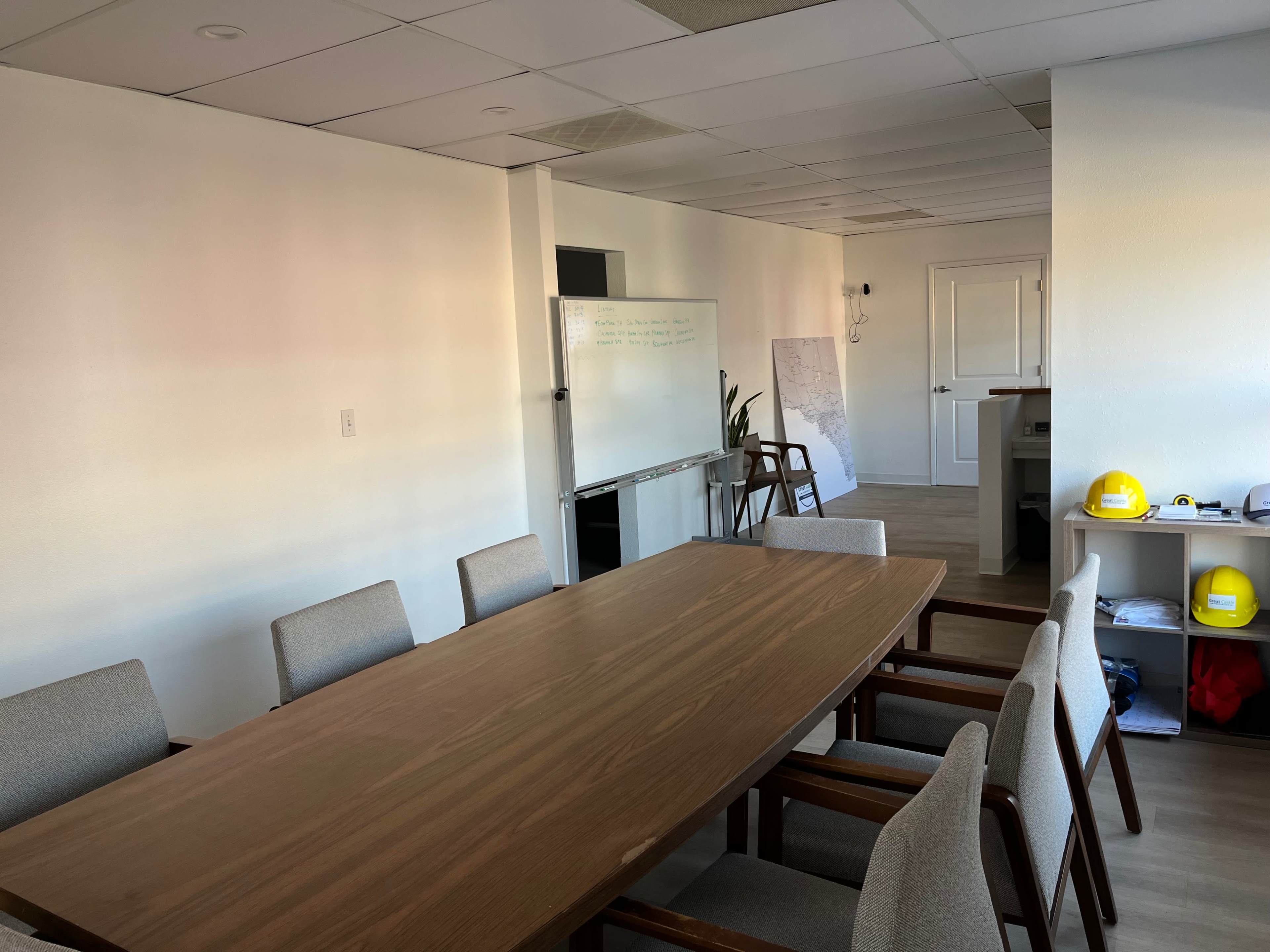 A conference room features a long wooden table surrounded by upholstered chairs, with whiteboards and construction helmets visible in the background.