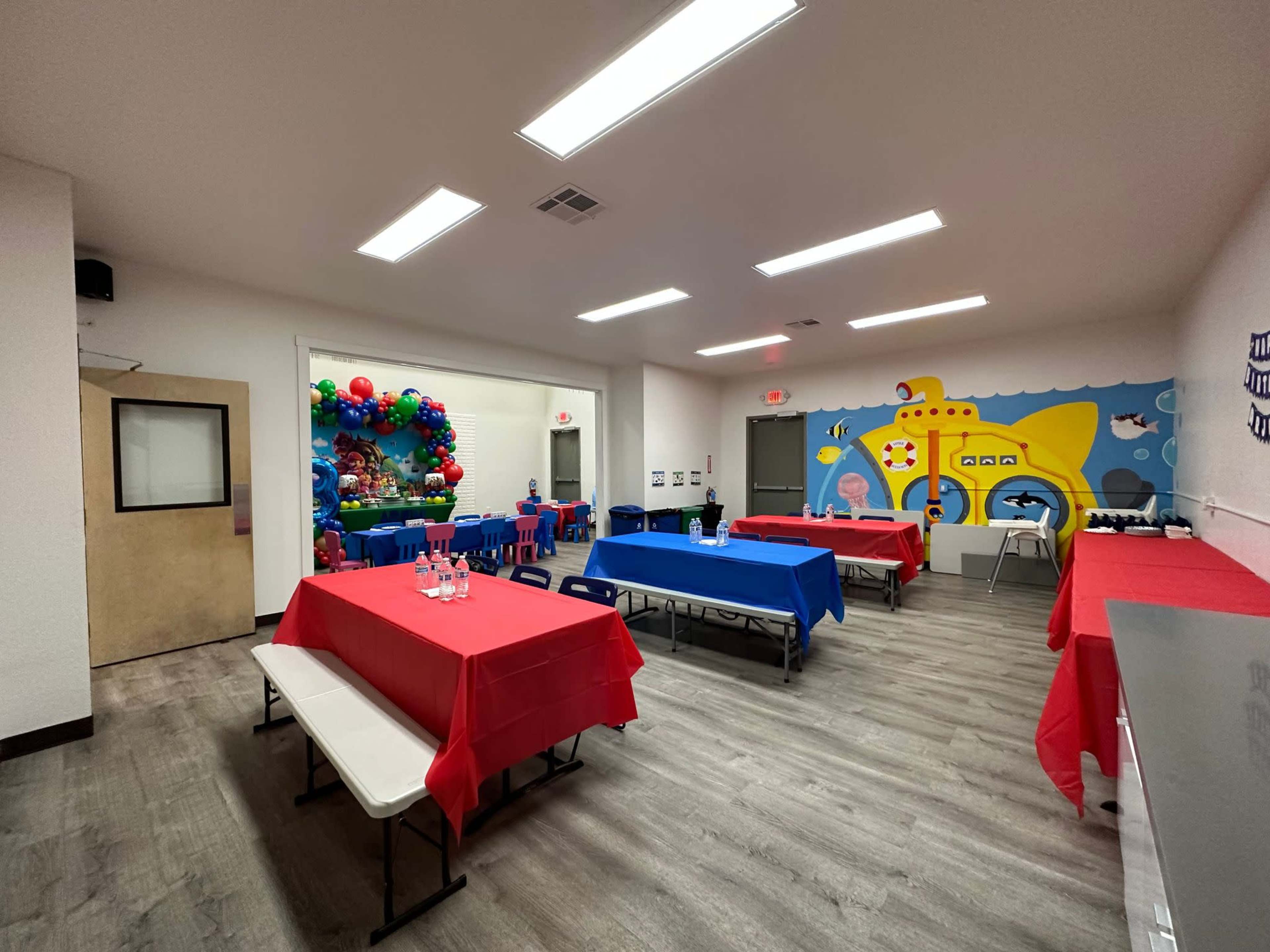 A brightly lit room features several tables covered in red and blue tablecloths, with a colorful balloon display and a mural of a submarine on one wall.