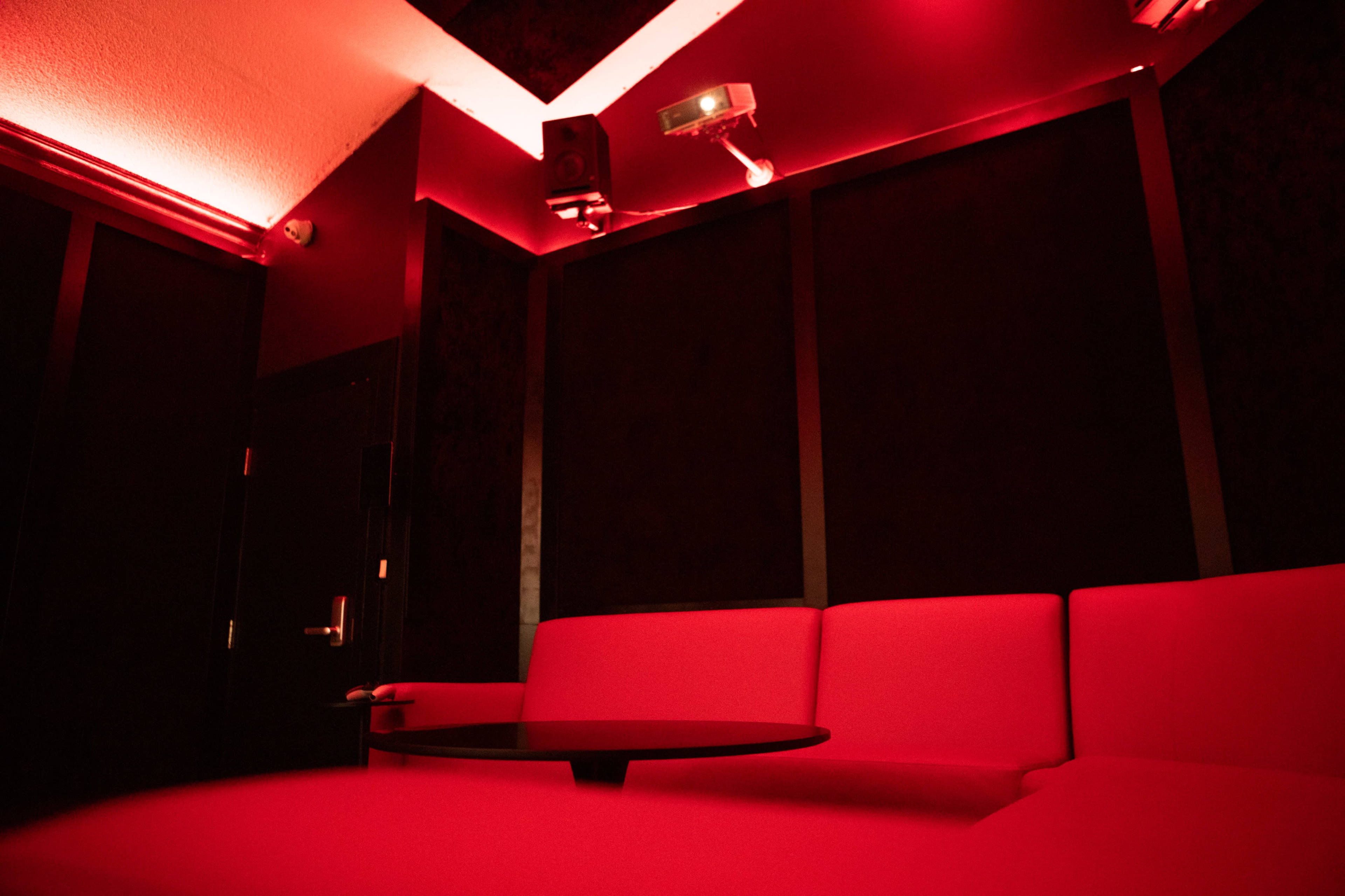 A small, dimly lit room features red walls and furniture, with a black table positioned in front of a red sectional couch.