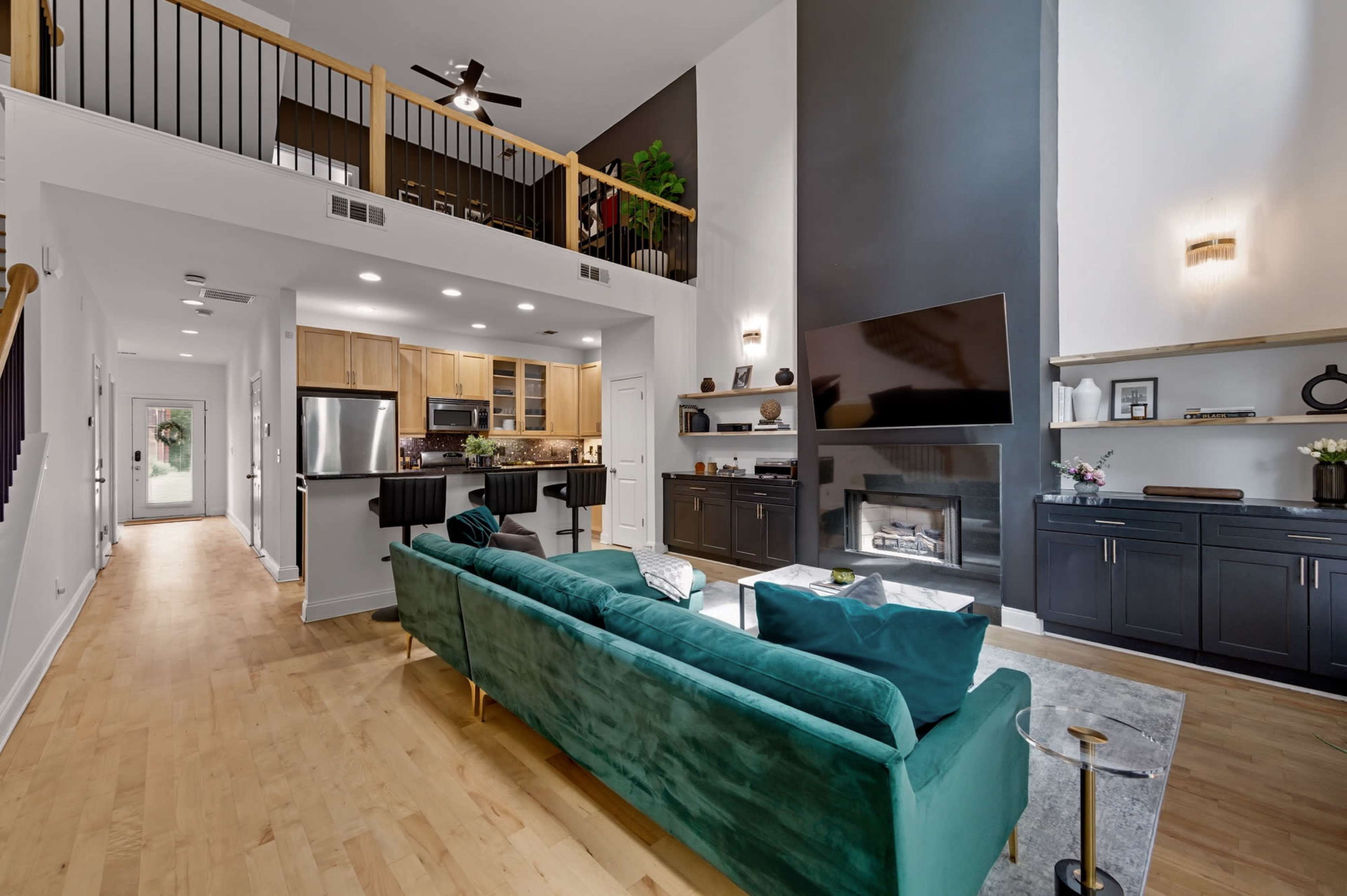 The image shows a modern, open-concept living area featuring a green velvet sofa, a mounted television, a fireplace, and a kitchen with wooden cabinets.
