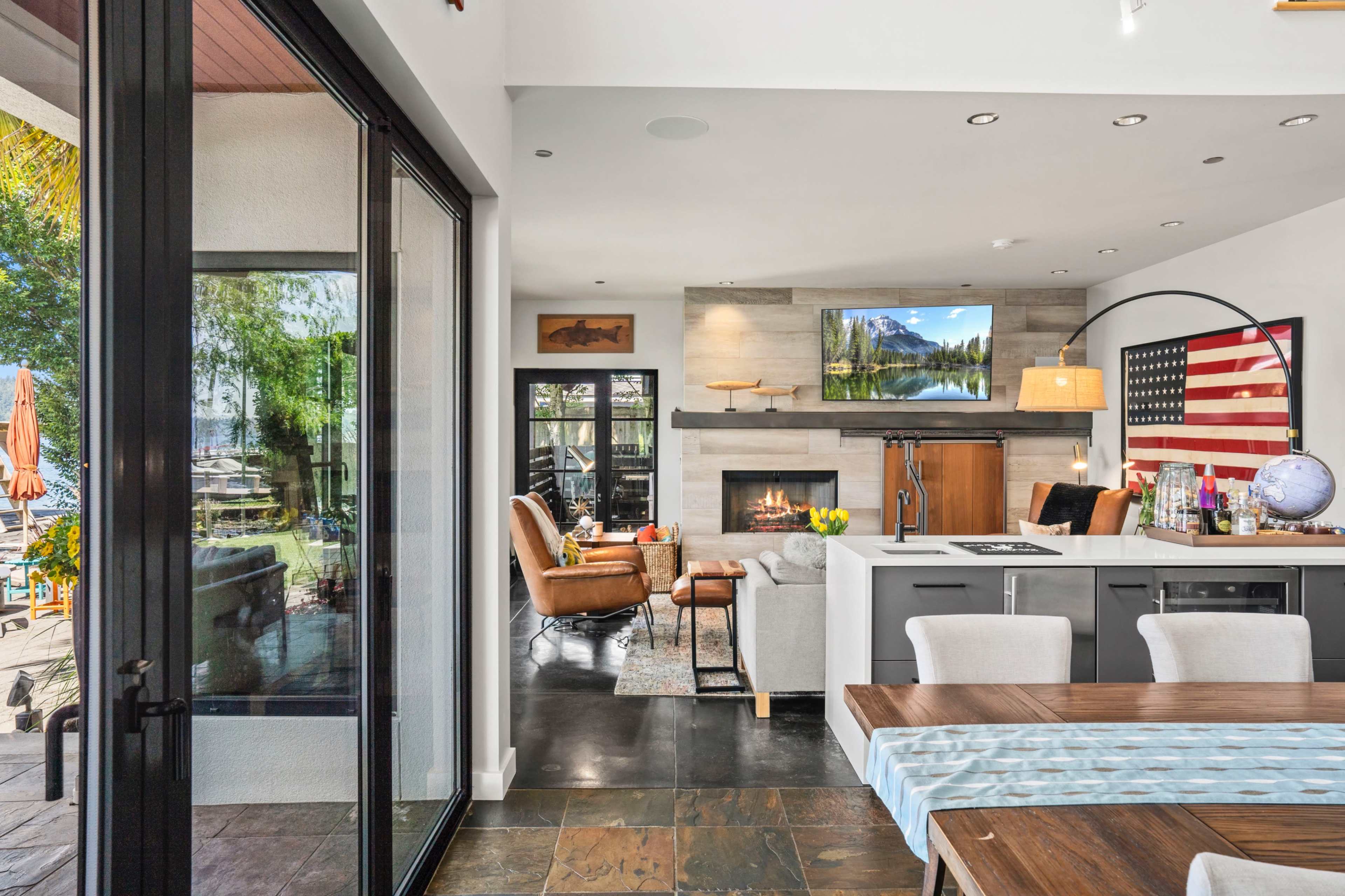 A modern open-concept living area featuring a kitchen, dining space, and sitting area with a large window overlooking a patio and waterfront.