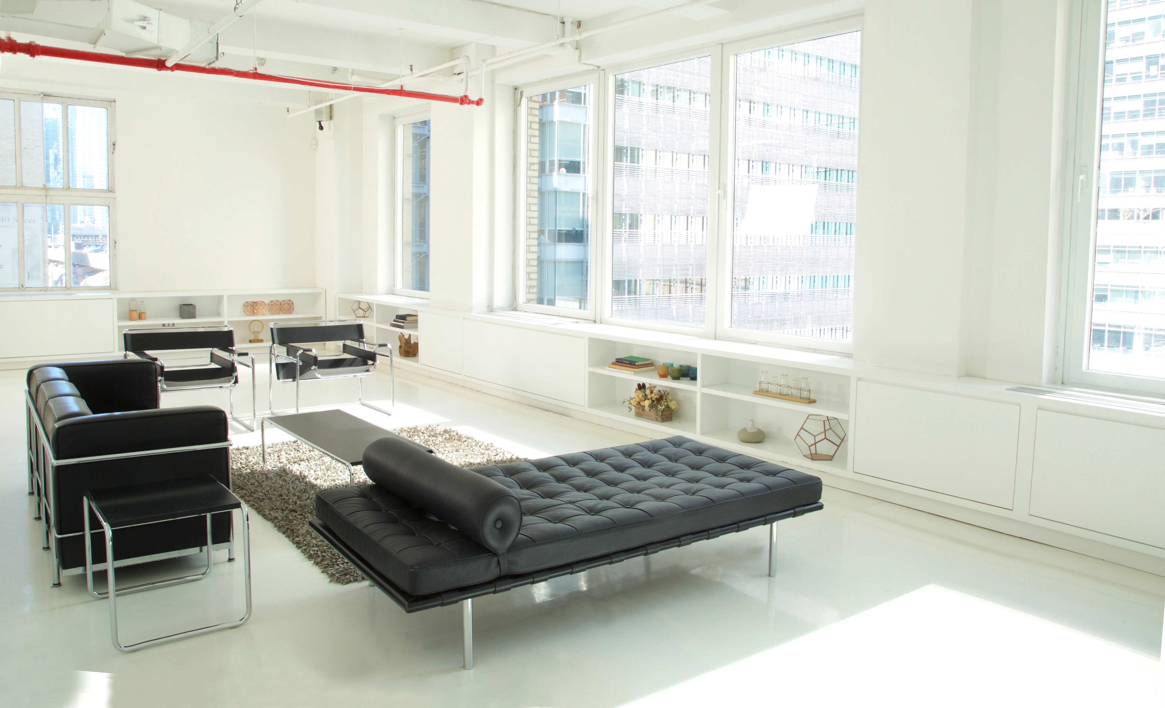 A modern, minimalist living space with large windows, a black sofa, and sleek furniture arrangements on a polished white floor.