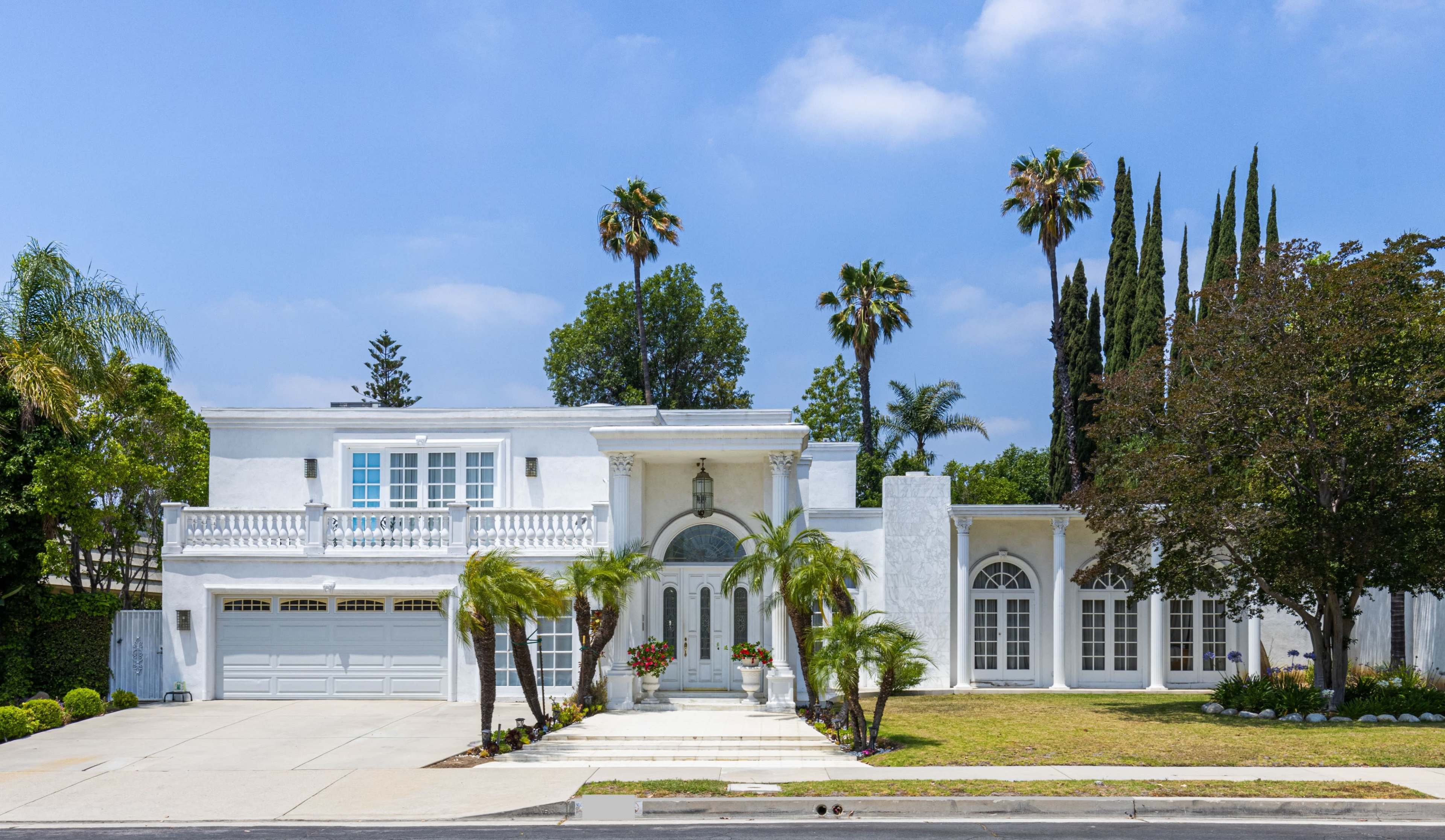 The image shows a large white house with a landscaped front yard, palm trees, and a two-car garage on a clear day.