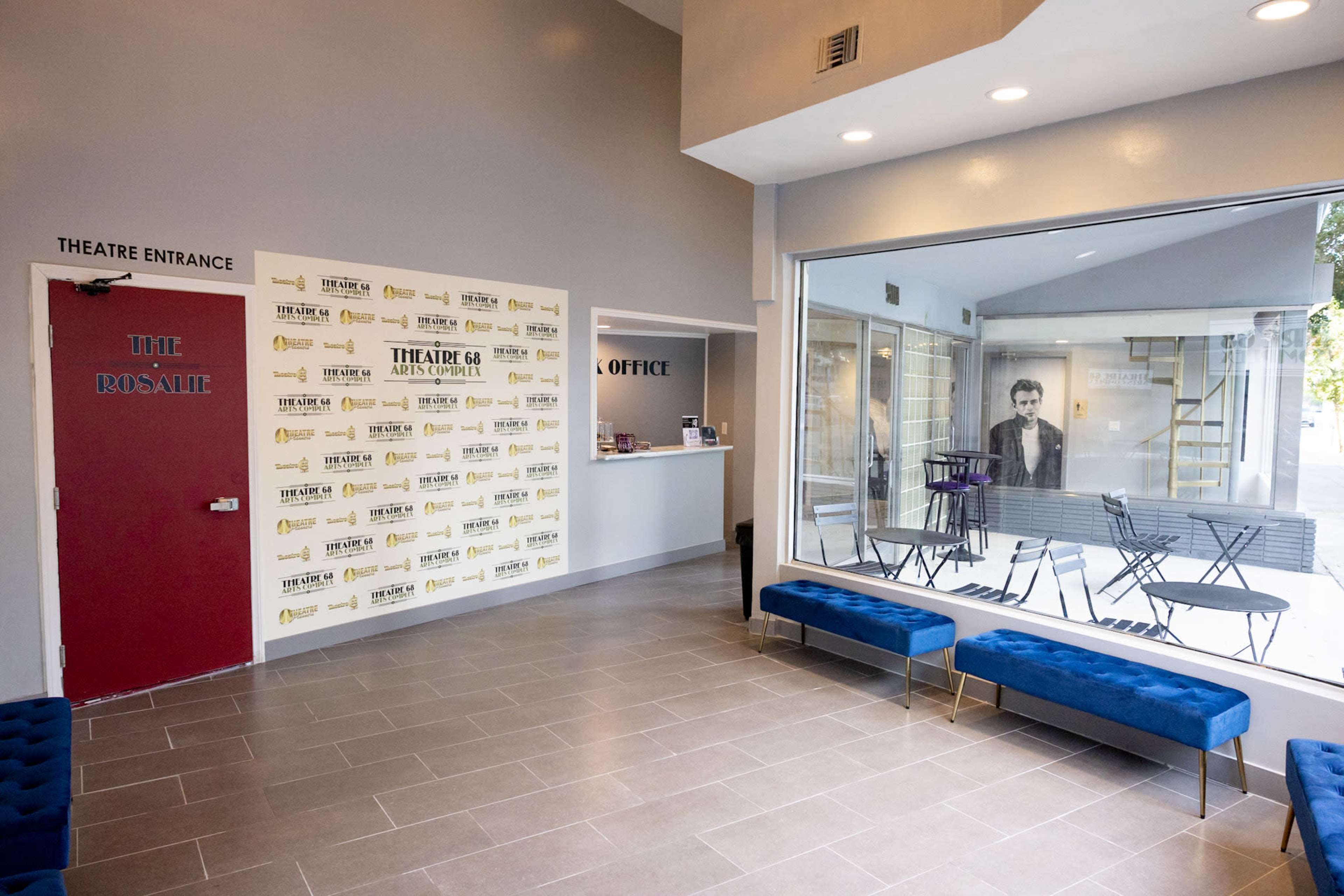 A theater lobby with a red door marked "THE ROSE," a wall covered in promotional posters, and a glass enclosure featuring seating and a figure of a man.