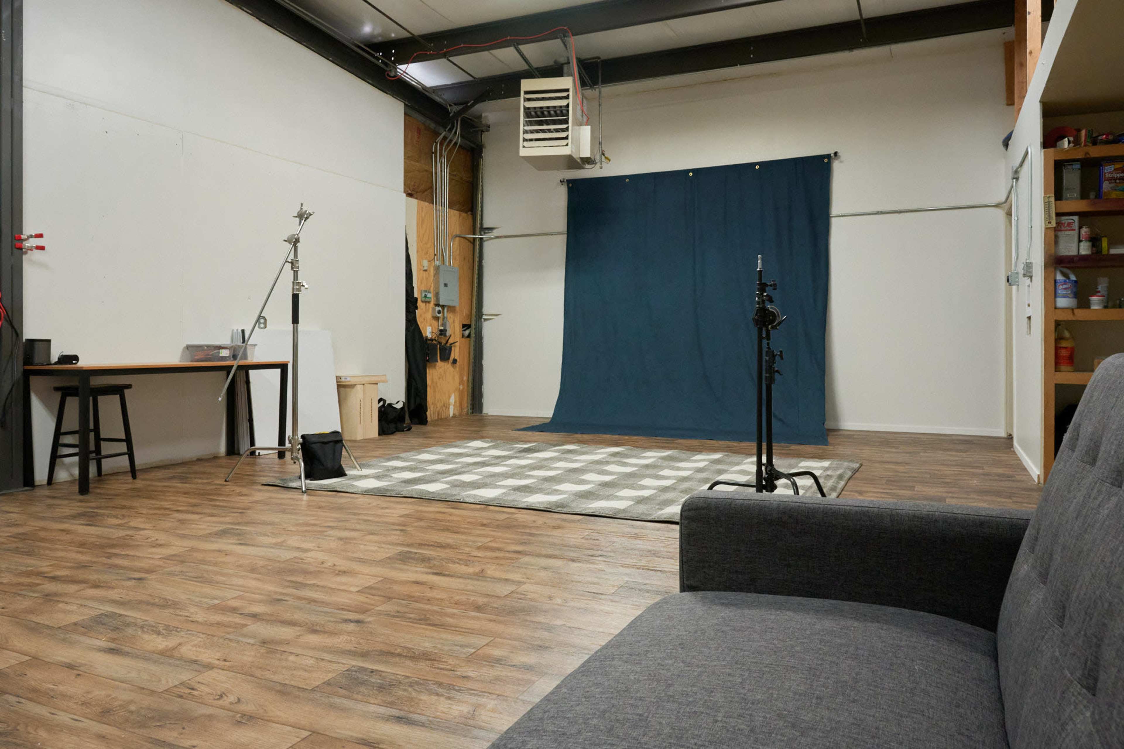 A spacious studio with a blue backdrop, a checkered rug, a tall stool, and a gray sofa.