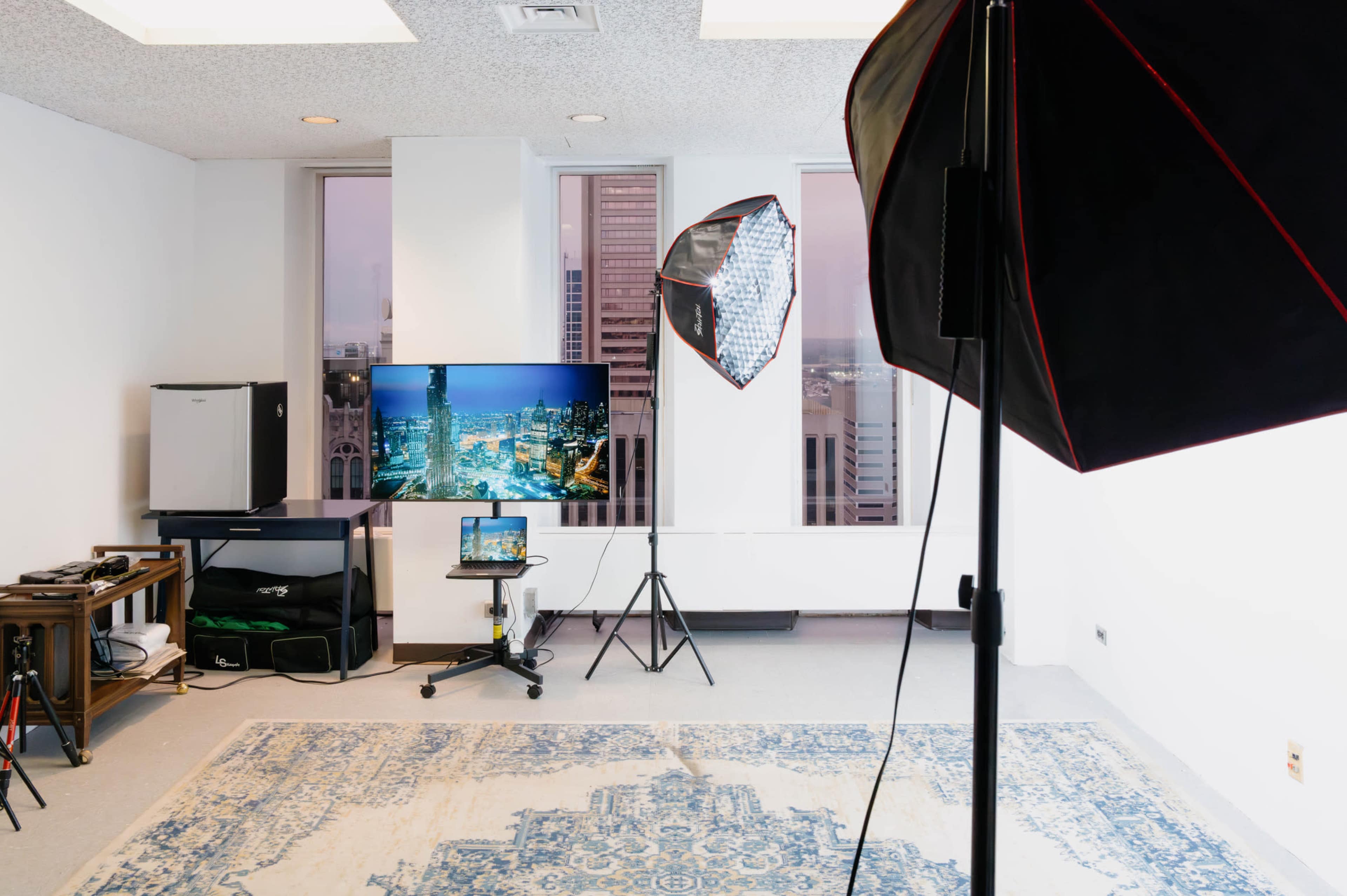 The image shows a minimalistic studio space featuring a television, a small table with a laptop, and two lighting setups on stands.