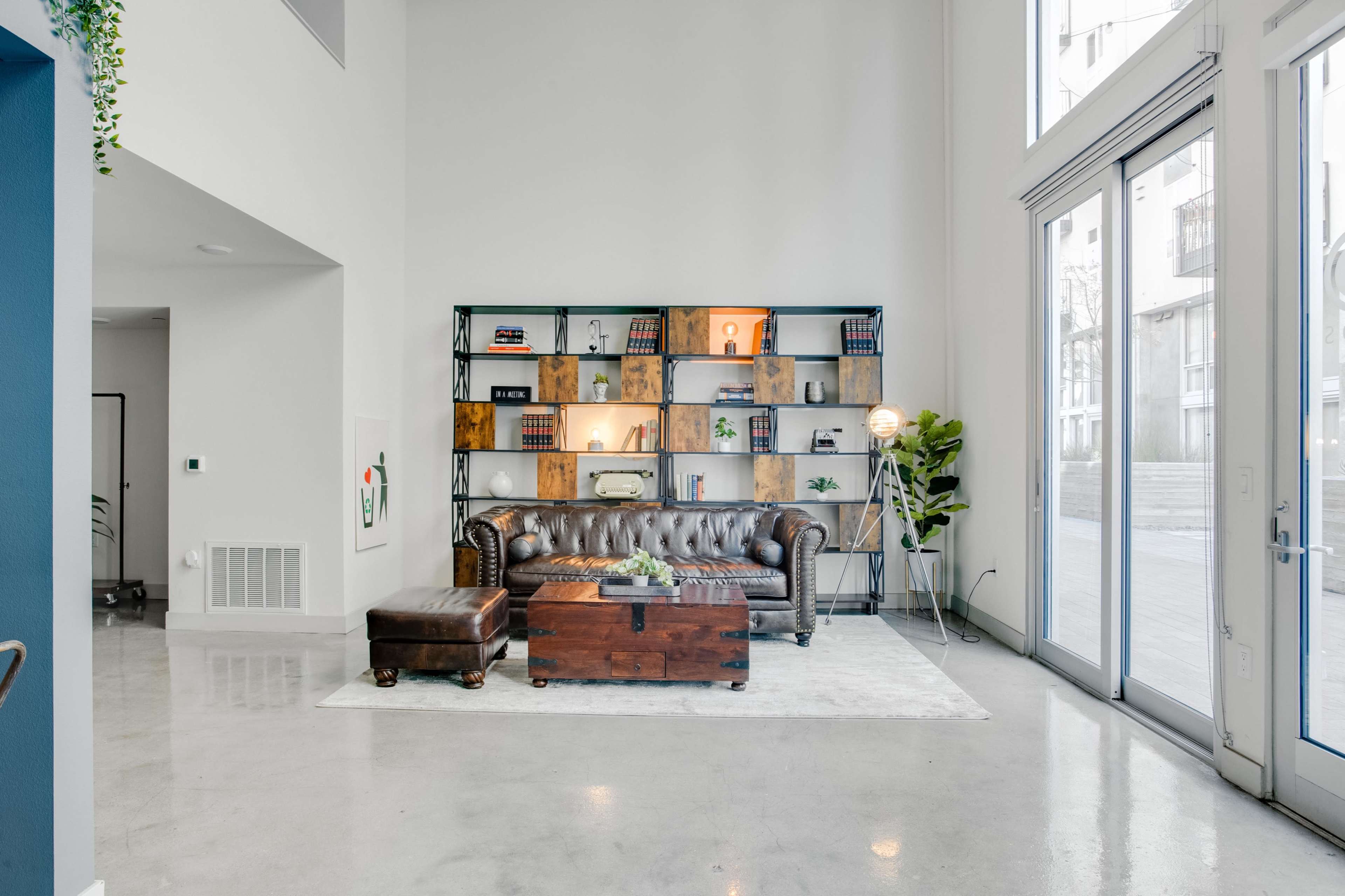 A leather sofa and ottoman sit on a rug in front of a wooden bookshelf filled with books and decorative items, in a spacious, well-lit room with large windows.