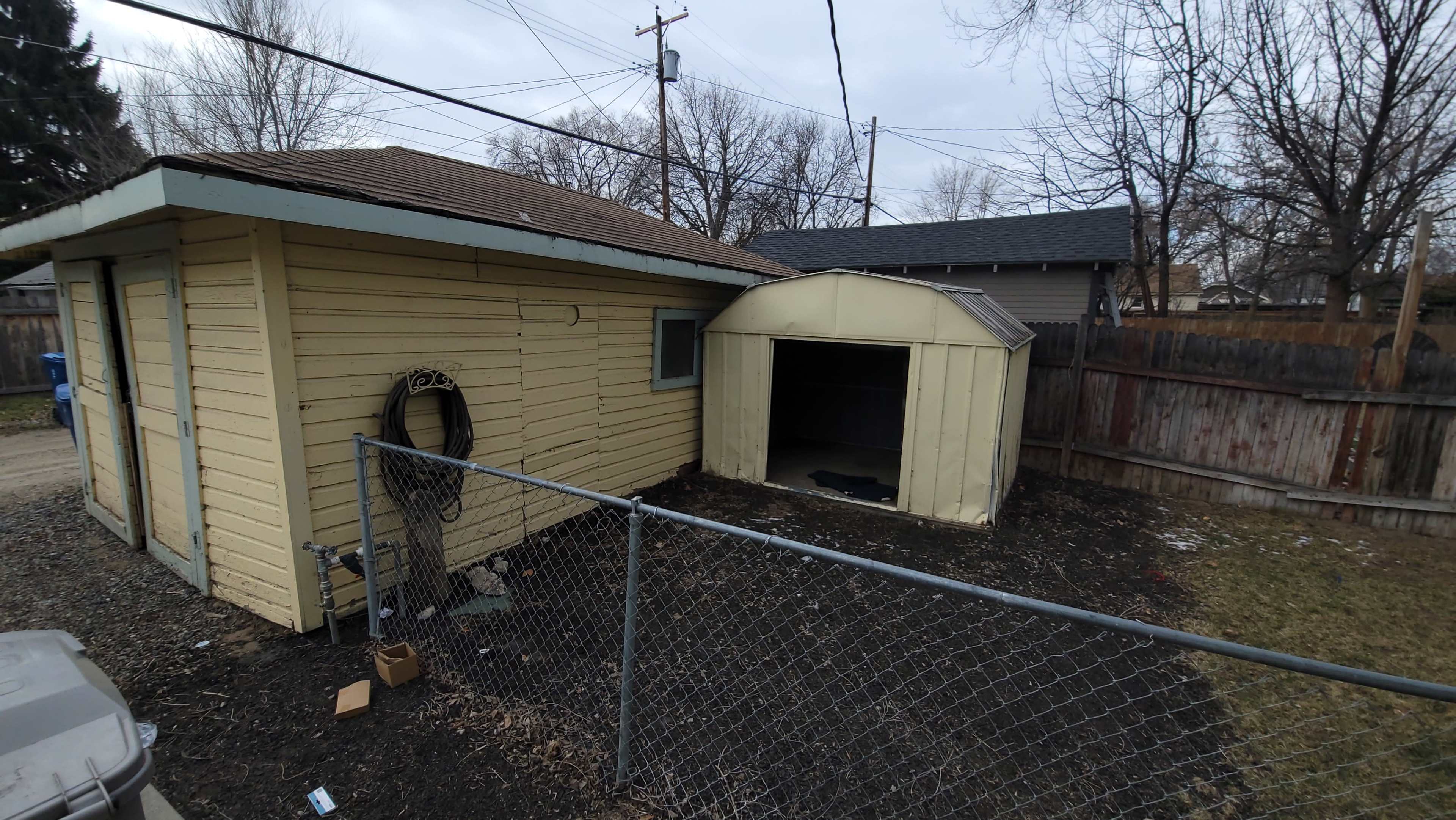 A yellow house with a brown roof sits next to a metal shed, enclosed by a chain-link fence in a yard with sparse grass and debris.