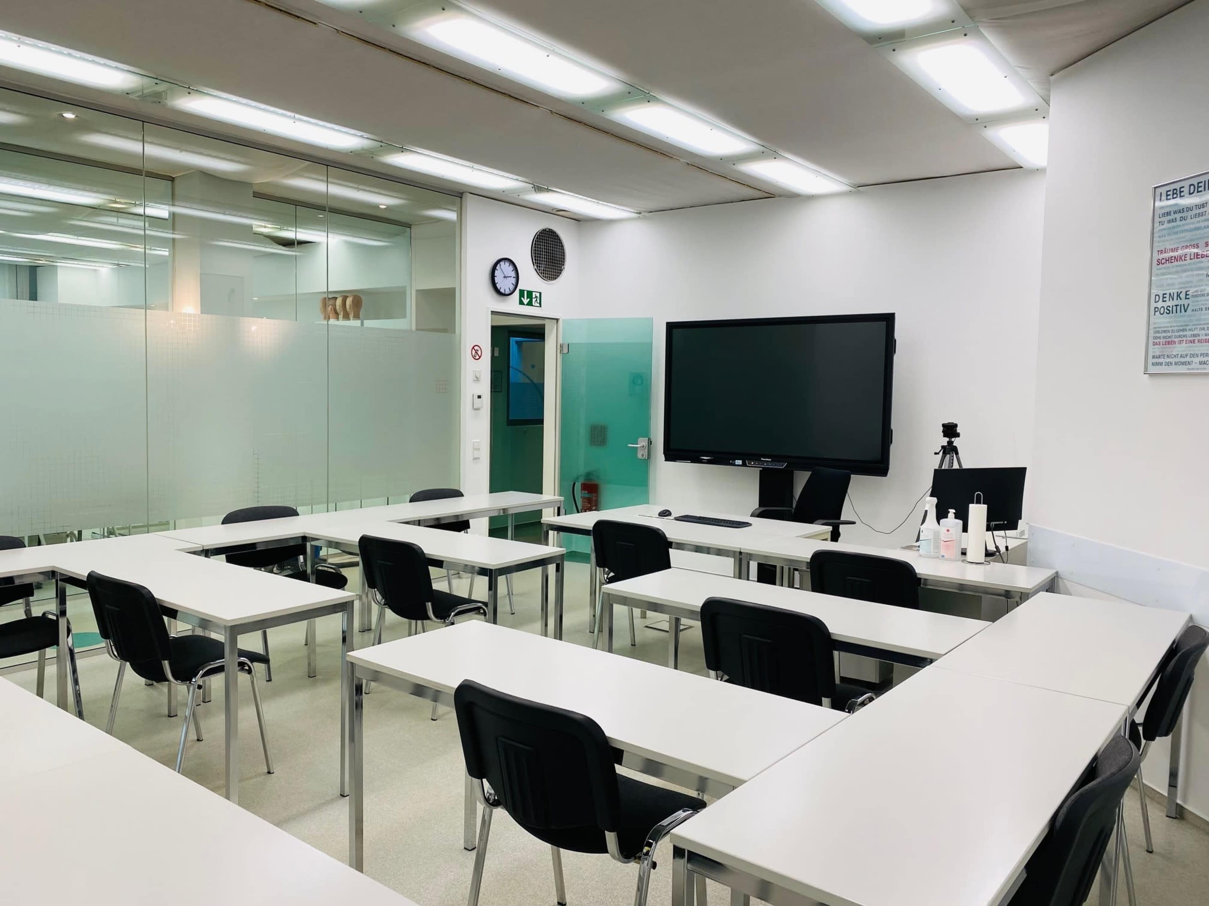 A modern classroom with rows of white tables and black chairs, equipped with a large screen, a clock, and glass walls.
