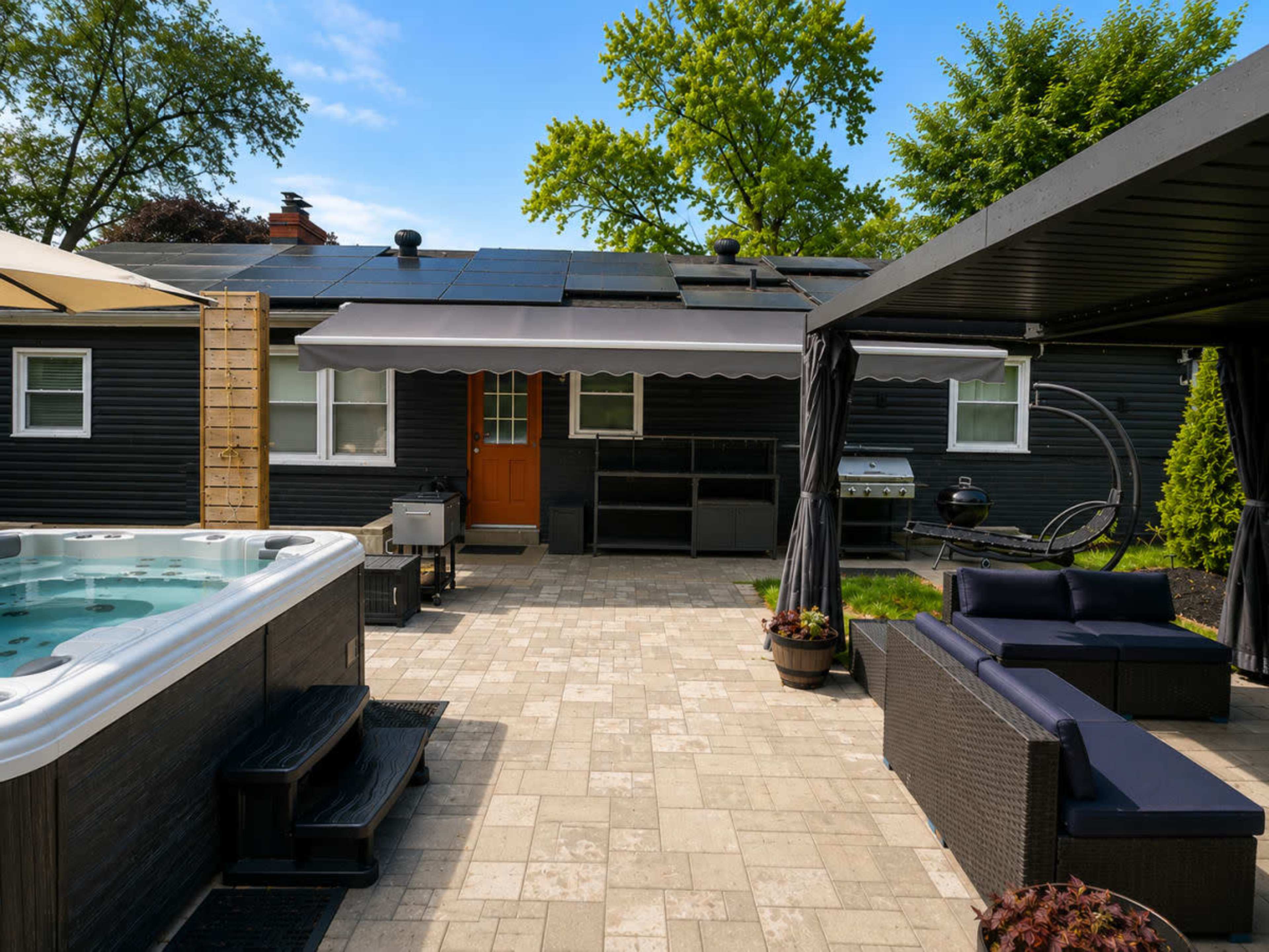 The image shows a backyard featuring a hot tub, seating area with outdoor furniture, a grill, and a house with solar panels on the roof.