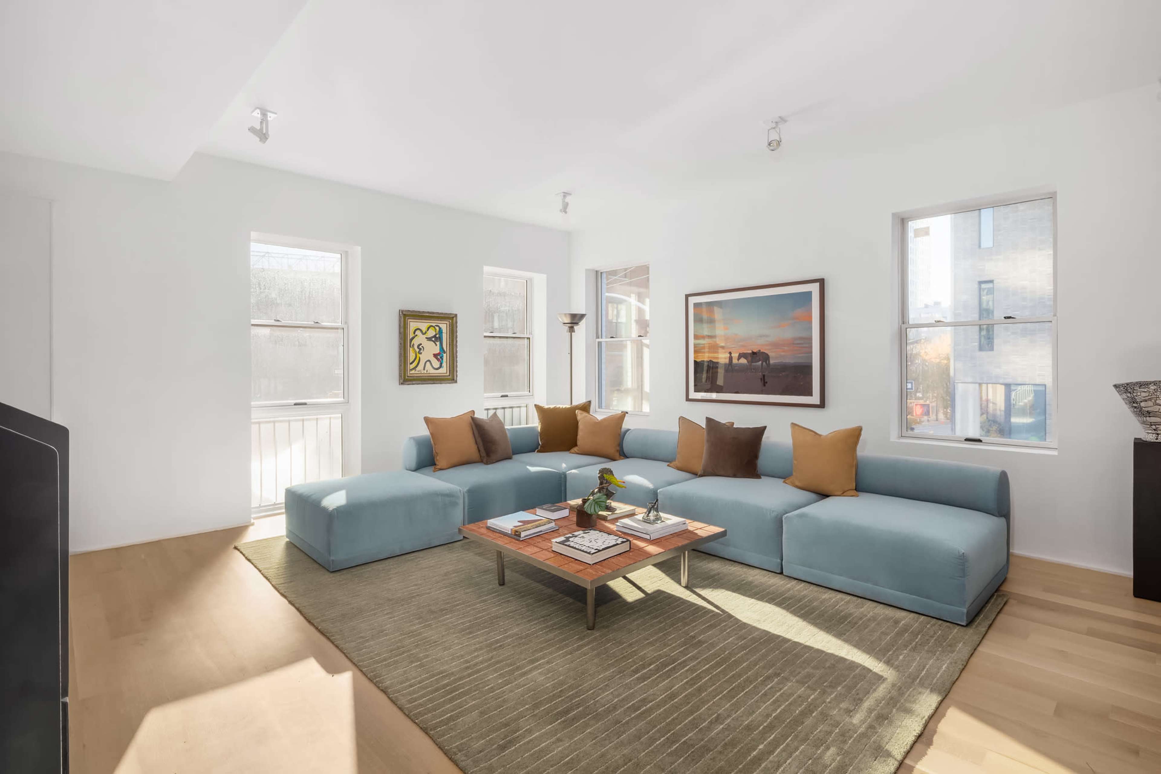The image shows a modern living room with a large light blue sectional sofa, a wooden coffee table, and several windows letting in natural light.
