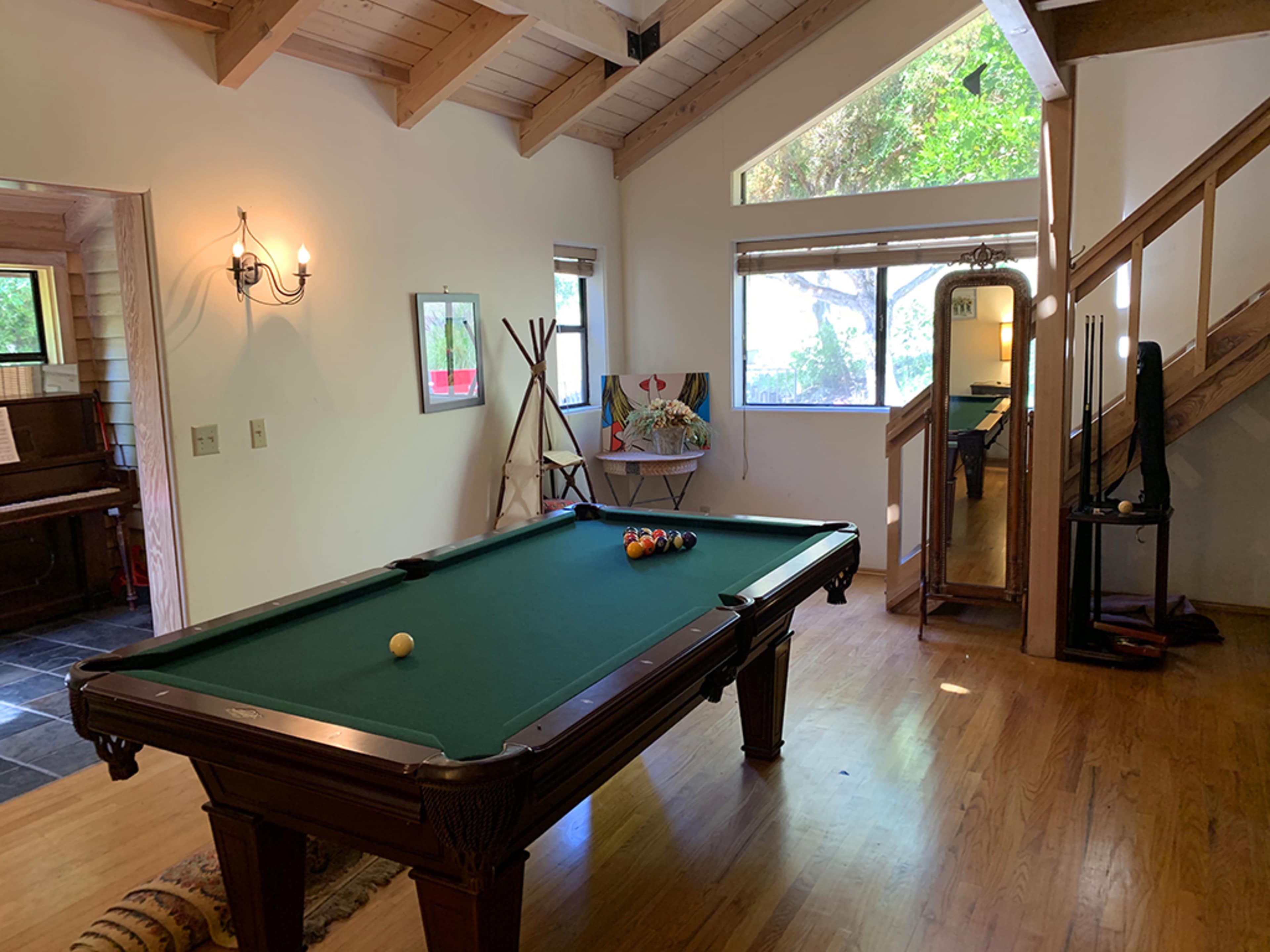 A spacious room featuring a green billiards table with a set of balls arranged on it, large windows letting in natural light, and a staircase leading to an upper level.