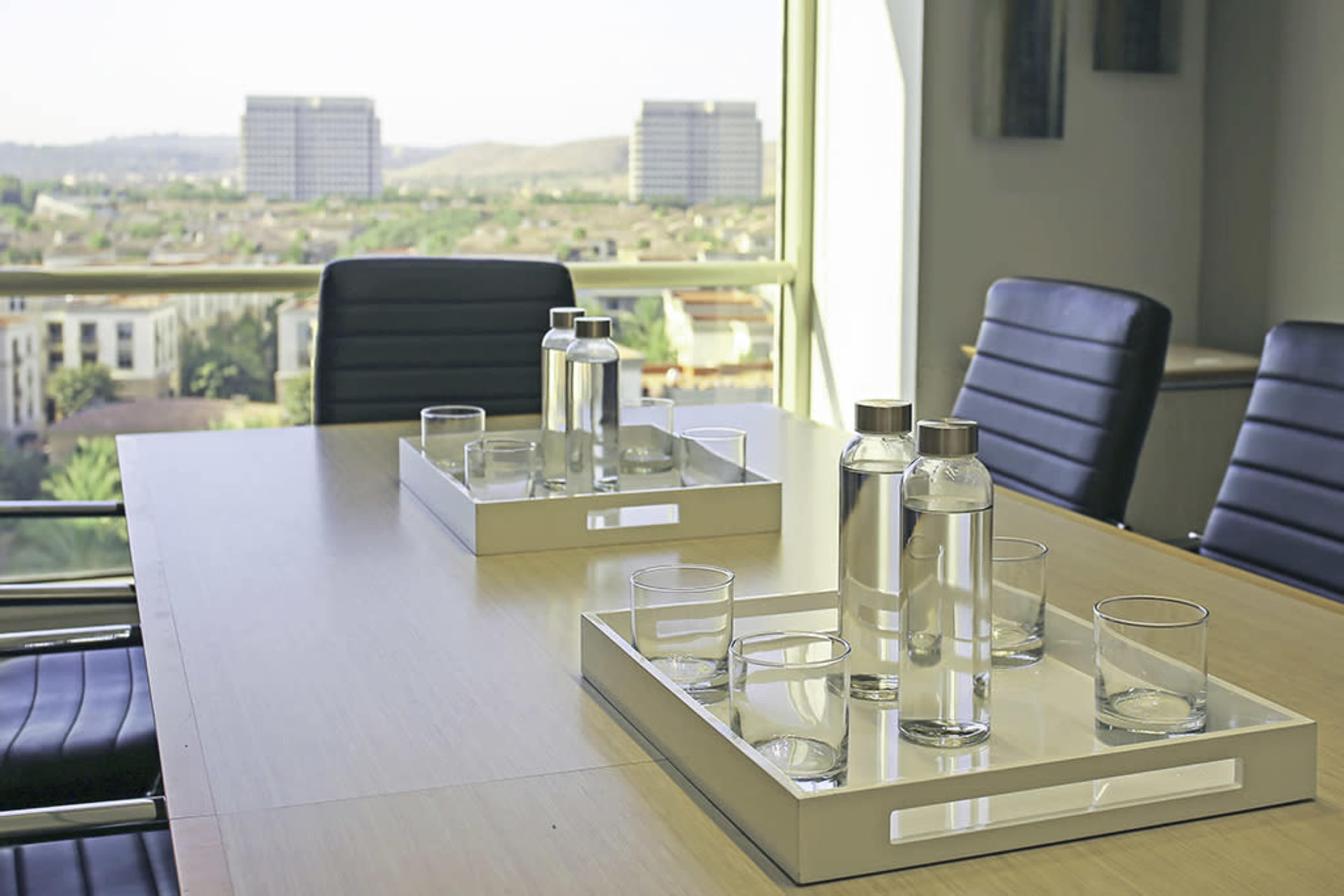 A conference room table with two trays of water bottles and glasses, overlooking a cityscape through large windows.