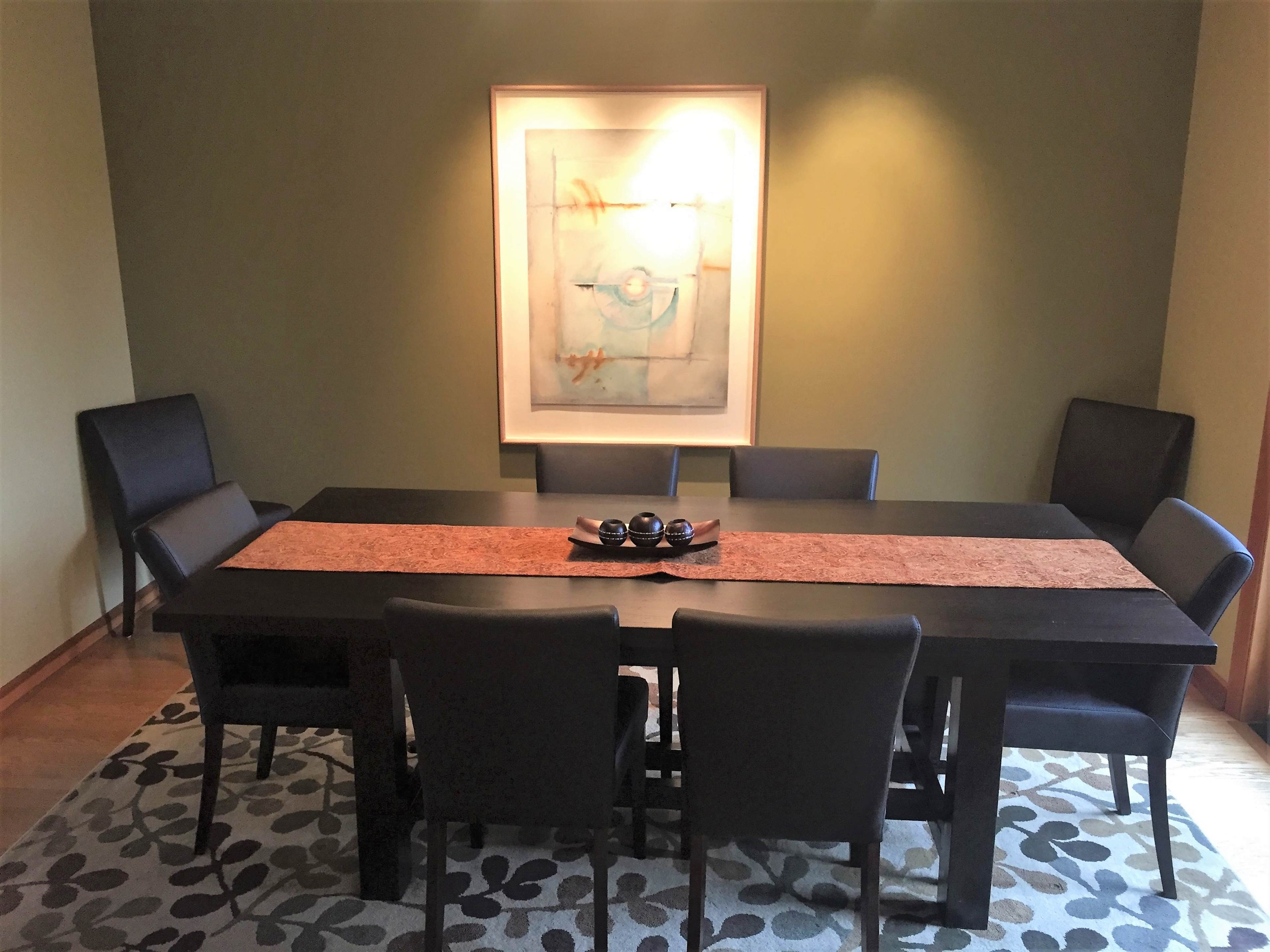 A dining room features a long black table surrounded by six chairs, with a decorative centerpiece and a painting on the wall.