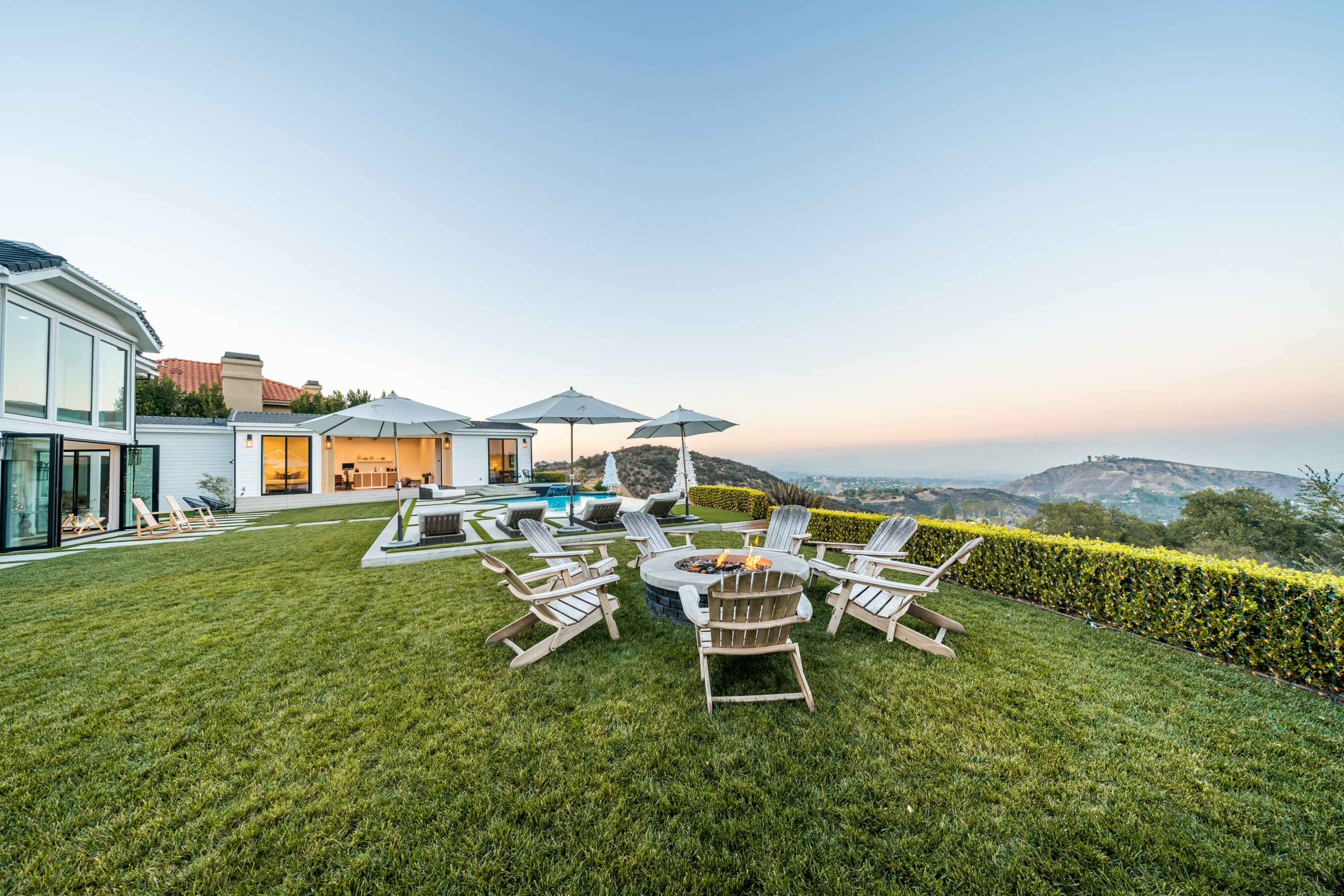 A modern hillside home features a well-maintained lawn with wooden chairs arranged around a fire pit, overlooking a scenic view at sunset.