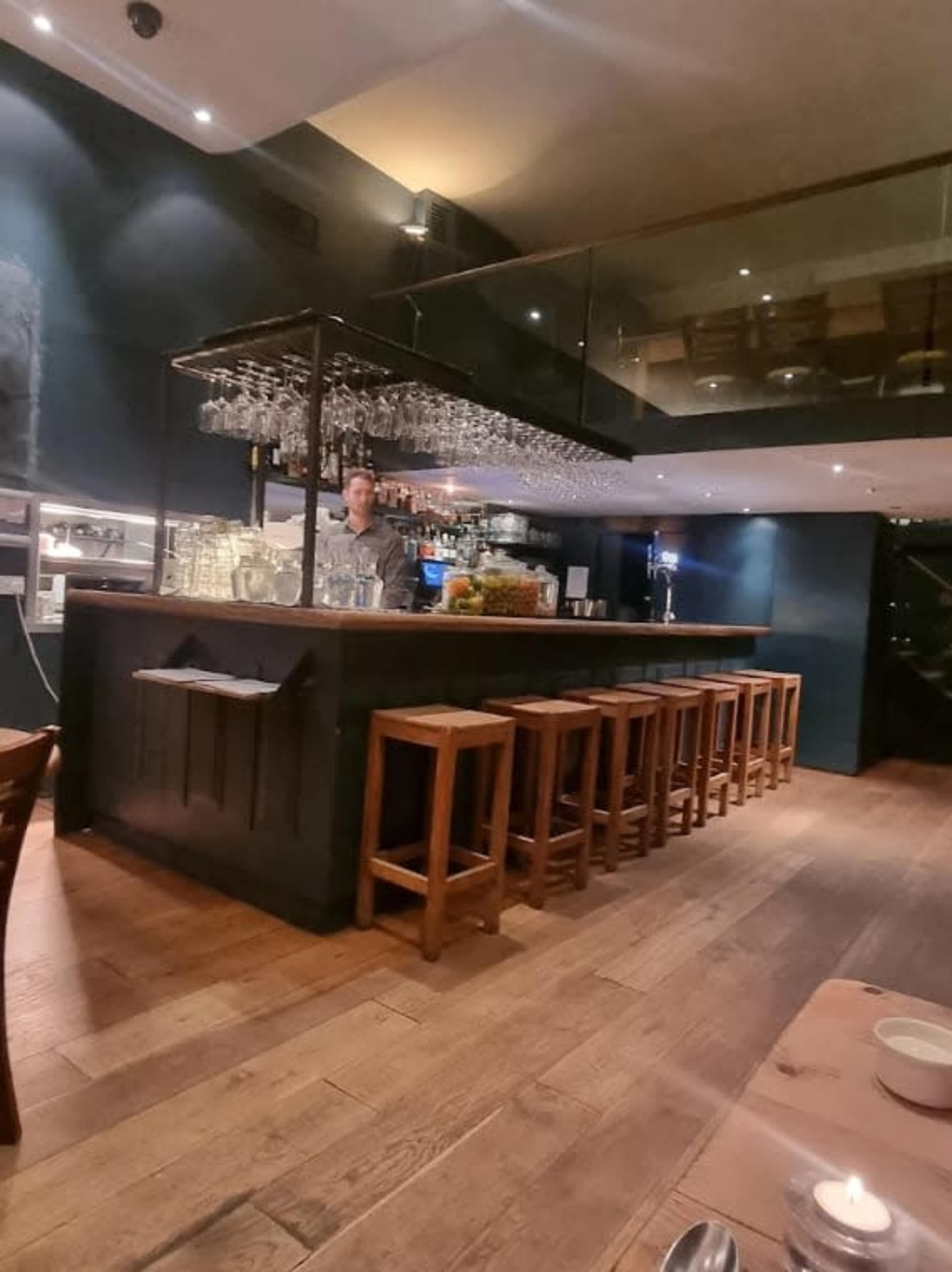 A bartender stands behind a wooden bar in a dimly lit restaurant, surrounded by shelves of glassware and a few tables in the foreground.