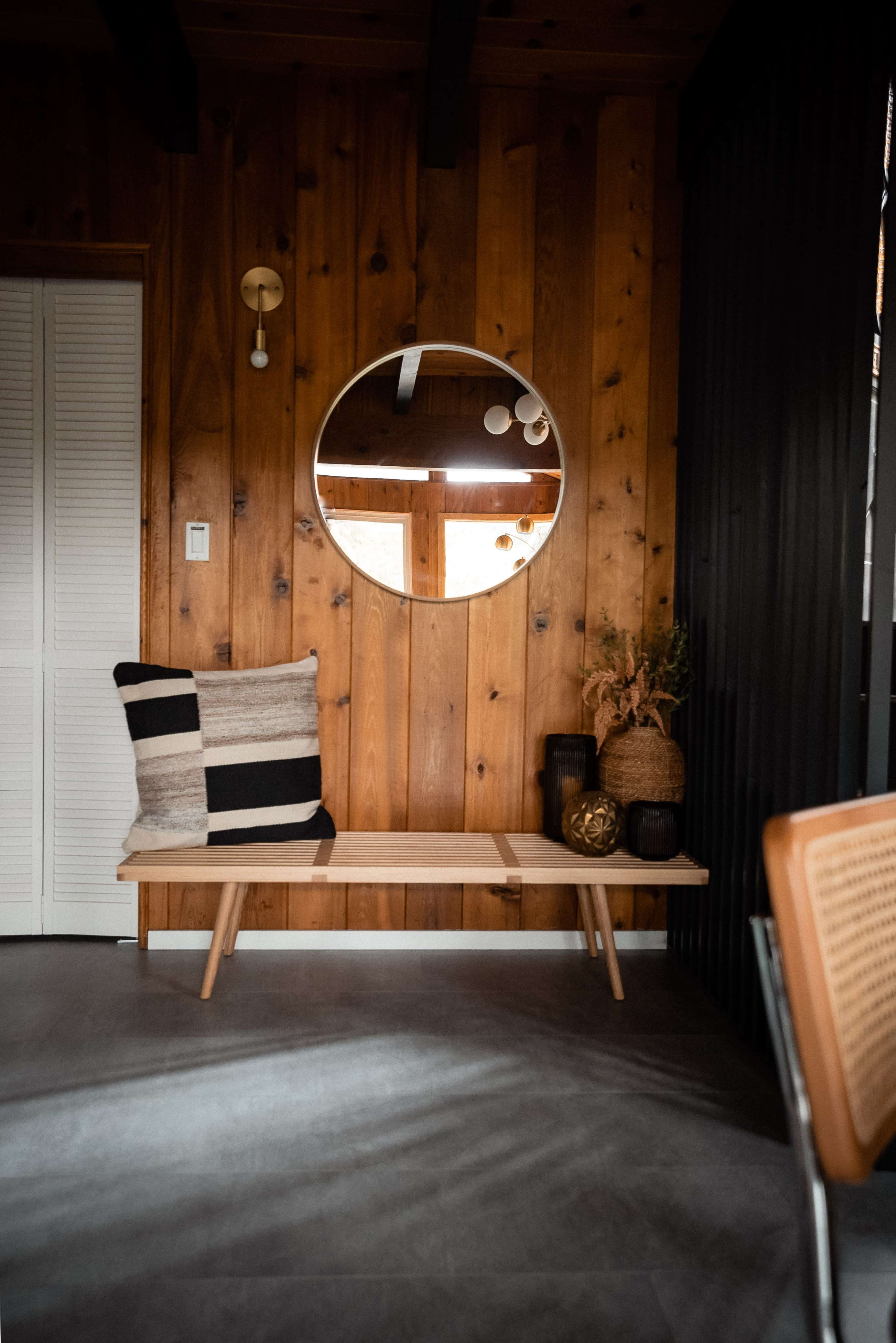 The scene features a wooden wall with a round mirror, a bench adorned with a striped pillow, and decorative items on display beside the bench.