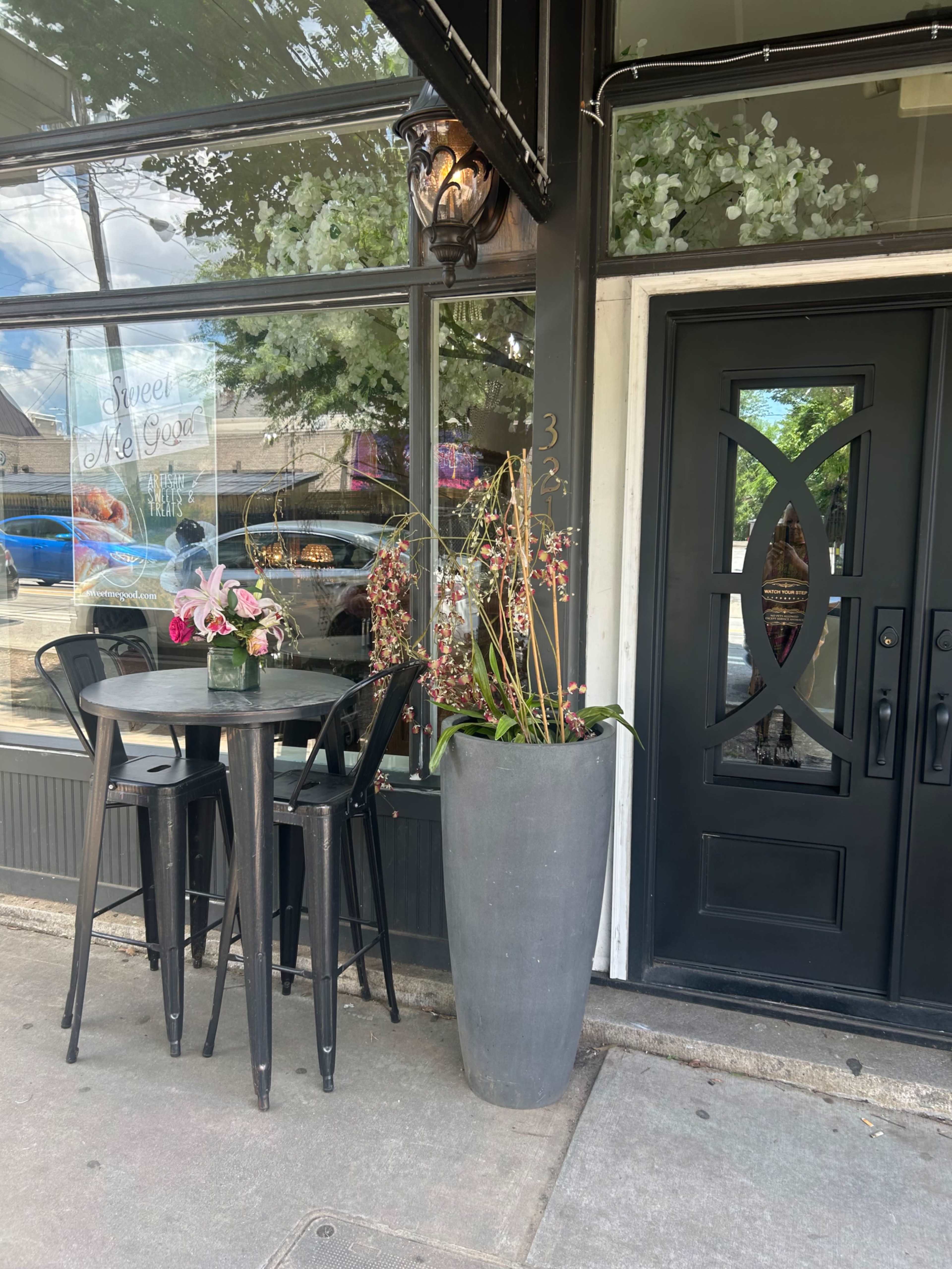 A small outdoor seating area with a glass table and black chairs is situated in front of a storefront featuring a large door and decorative potted plants.