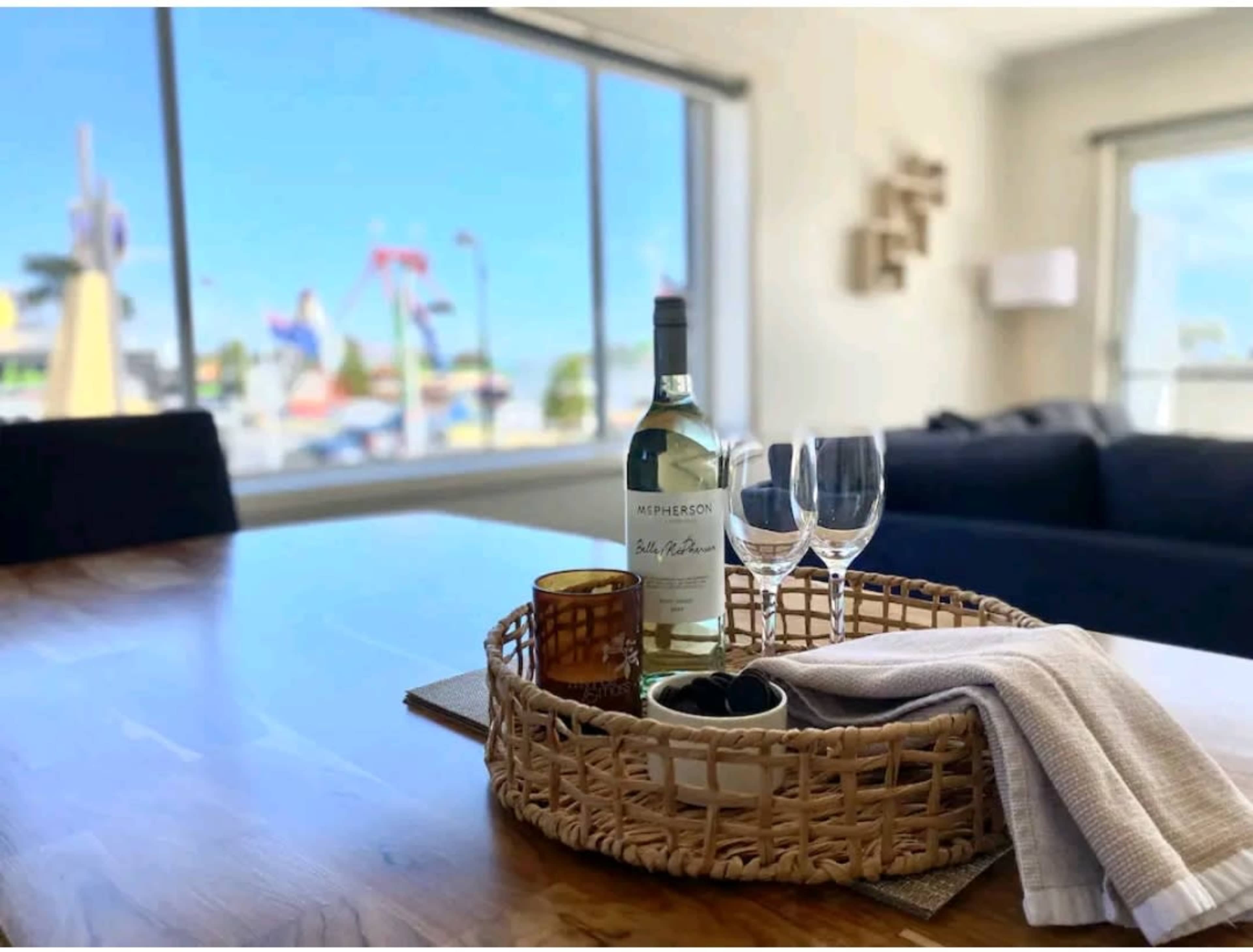 A bottle of white wine and two glasses rest on a woven tray on a wooden table, with a colorful amusement park visible through the window in the background.