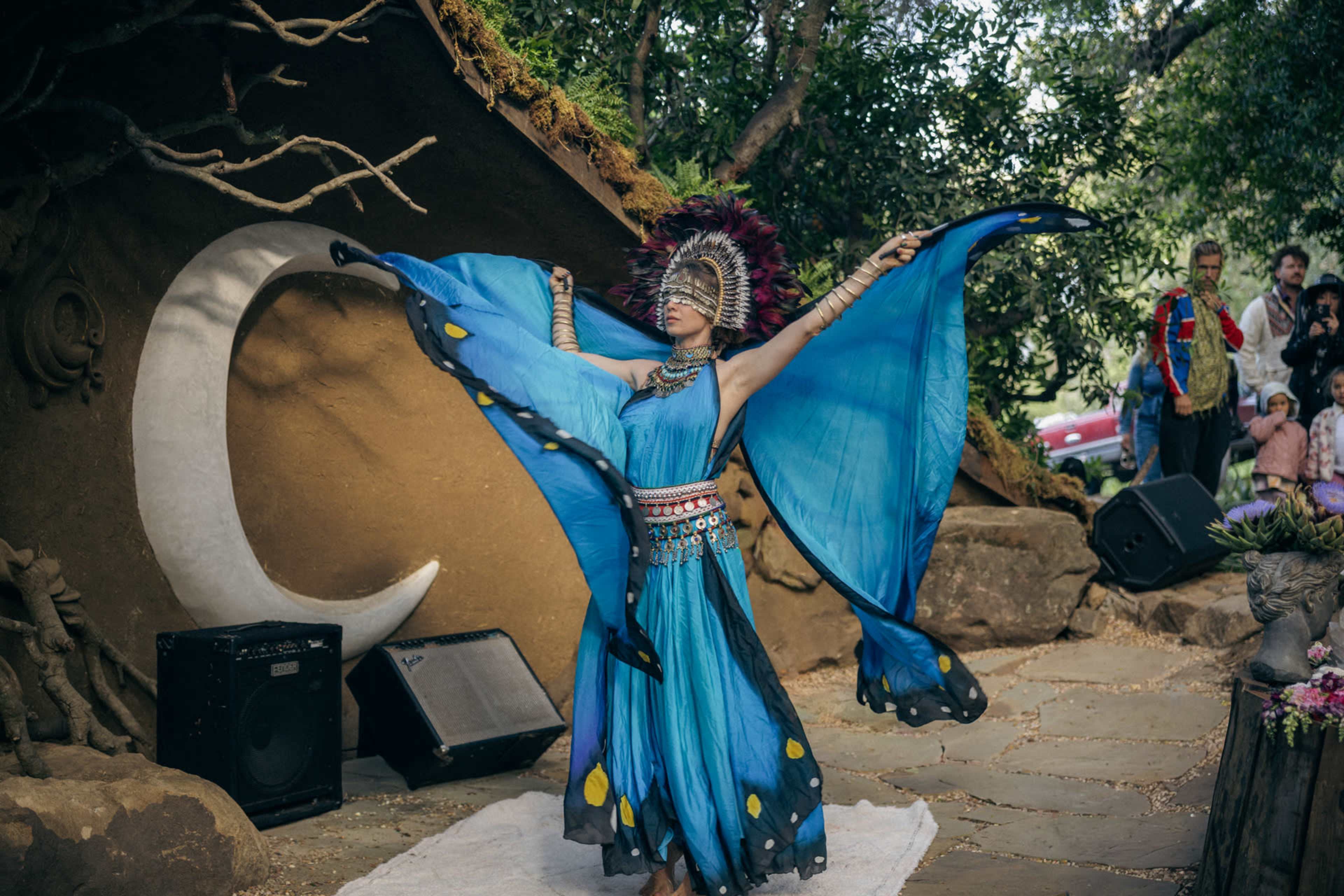 A dancer wearing a blue costume with large wing-like sleeves performs in front of a stone backdrop featuring a crescent moon.