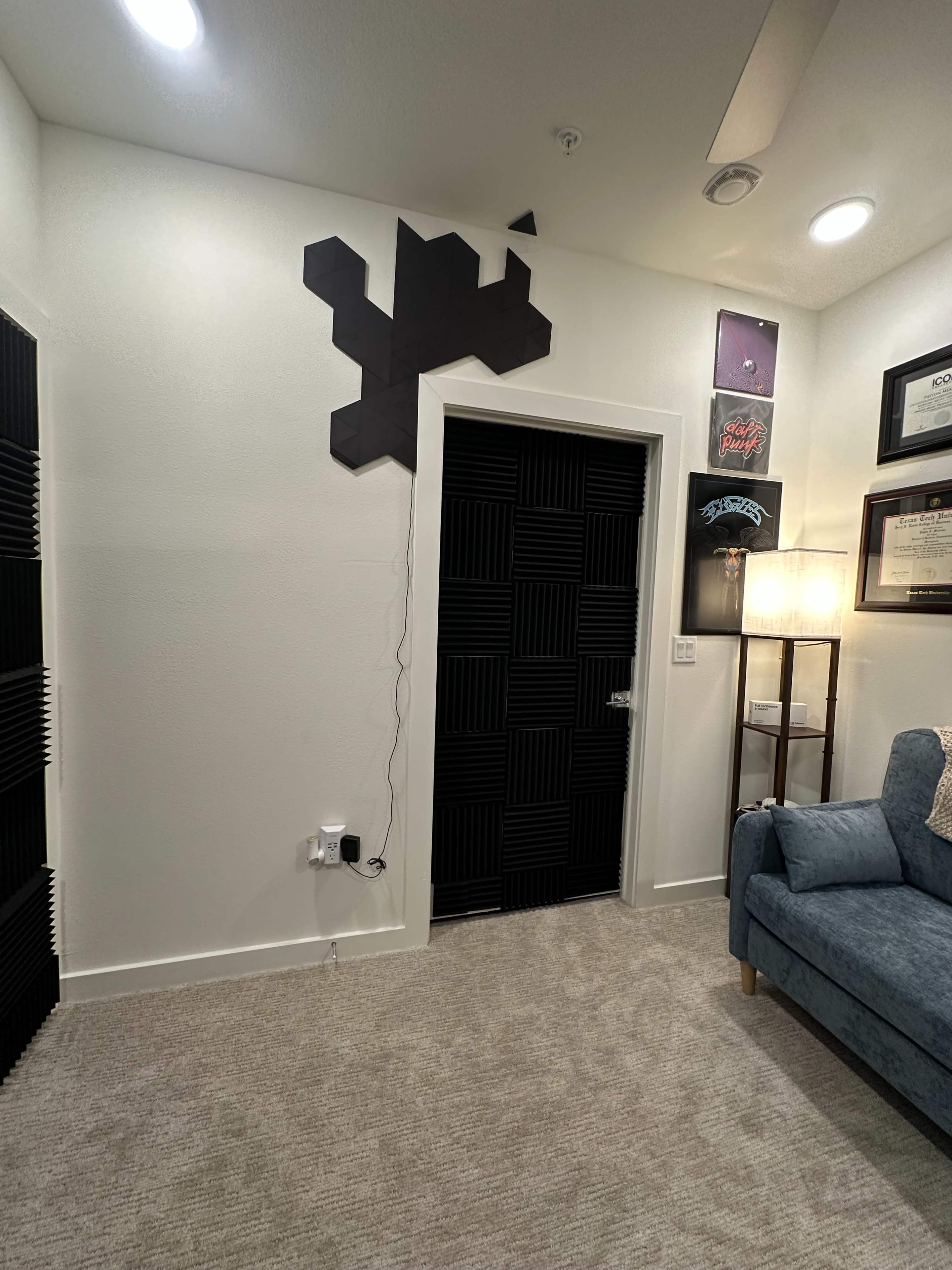 The image shows a corner of a room featuring black acoustic panels on the door, a light fixture on the ceiling, and a blue couch against the wall.