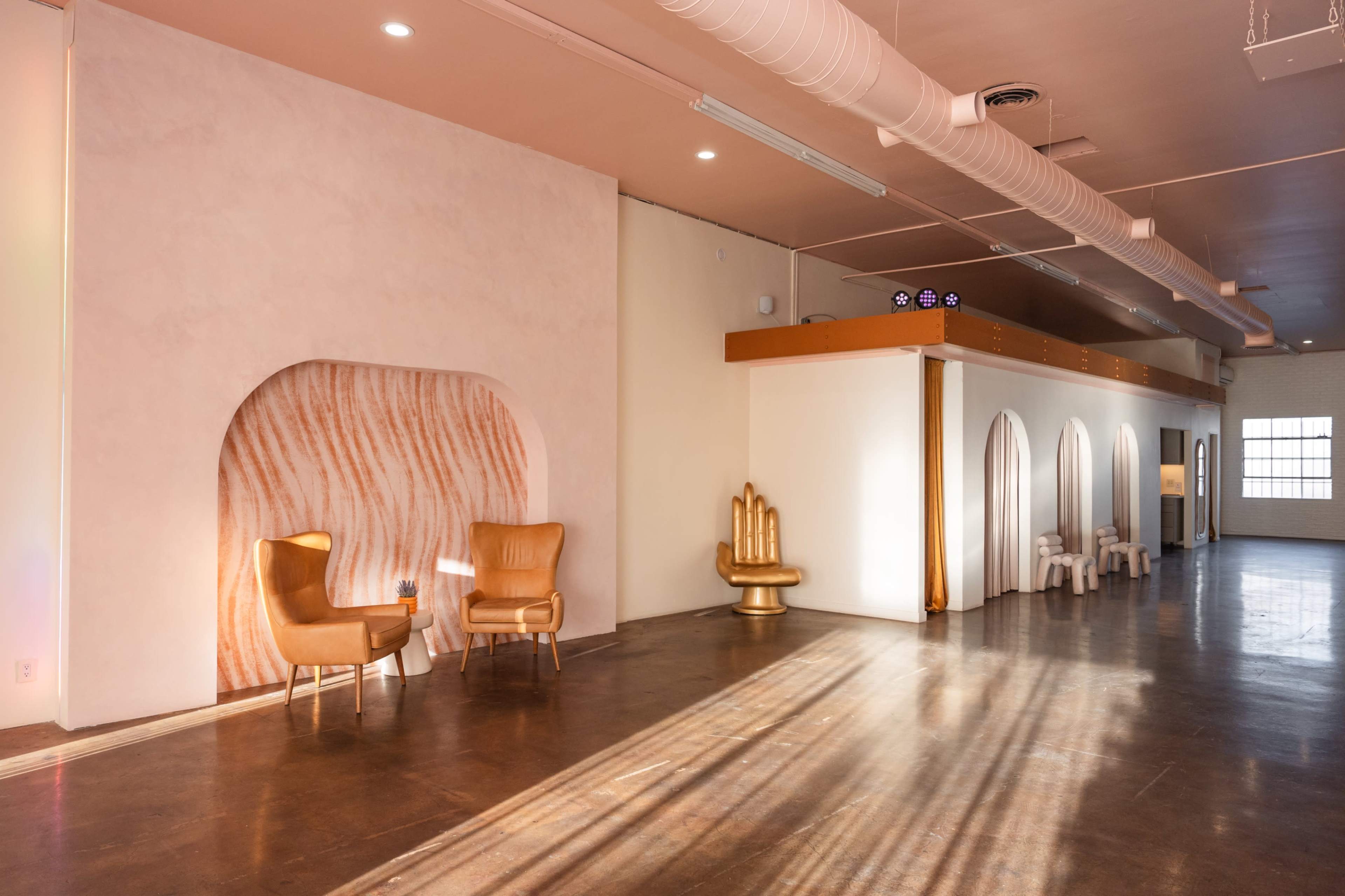 A spacious interior features two golden chairs positioned against a textured wall, with arched openings in a white partition and ample natural light streaming through large windows.