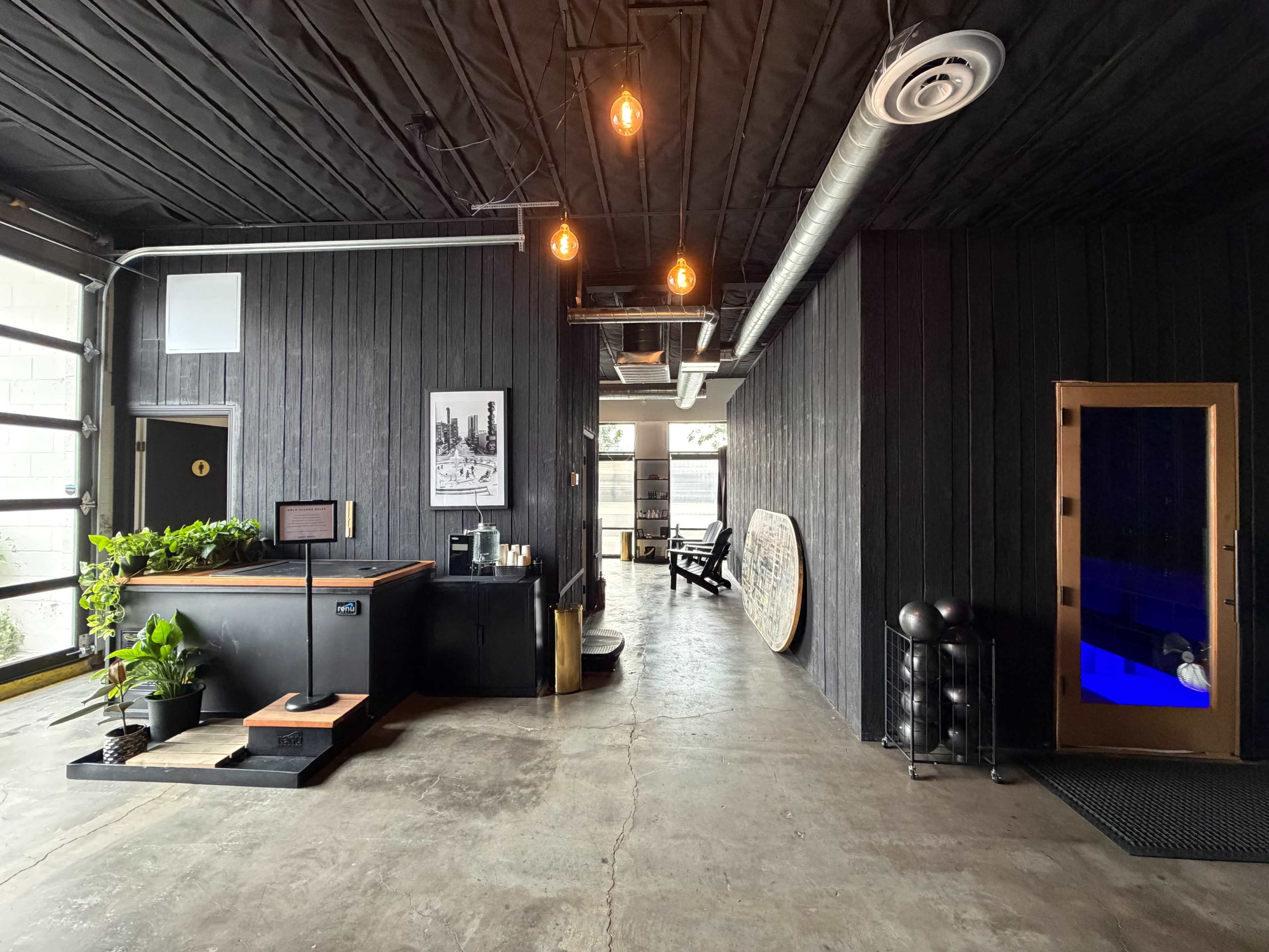 The image shows a modern interior space with a reception area featuring a counter, plants, and a sauna door next to a hallway leading to additional seating.