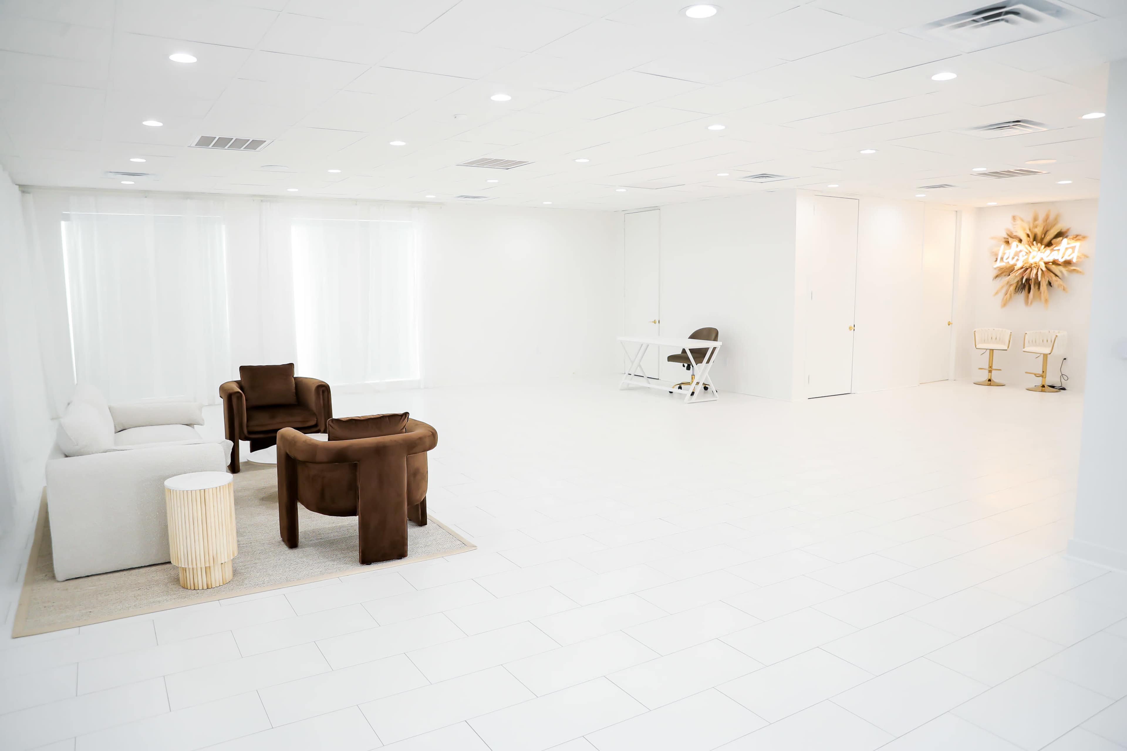 A spacious, well-lit room with white walls and floor, featuring a light-colored sofa, two brown armchairs, a small side table, and minimal furniture against a backdrop of large windows.