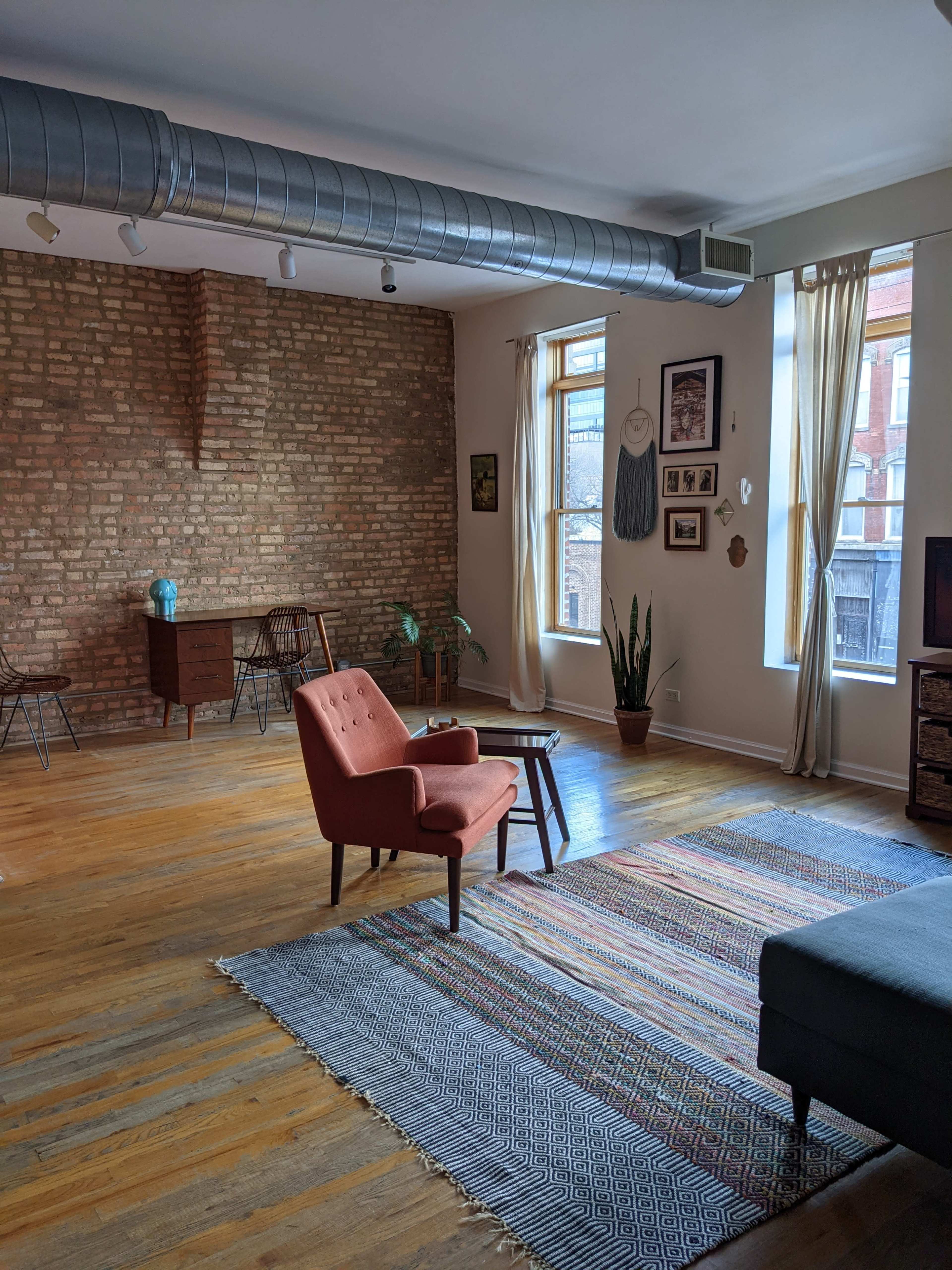 The image shows a spacious living room with exposed brick walls, hardwood flooring, a large area rug, and a mix of vintage and modern furniture.