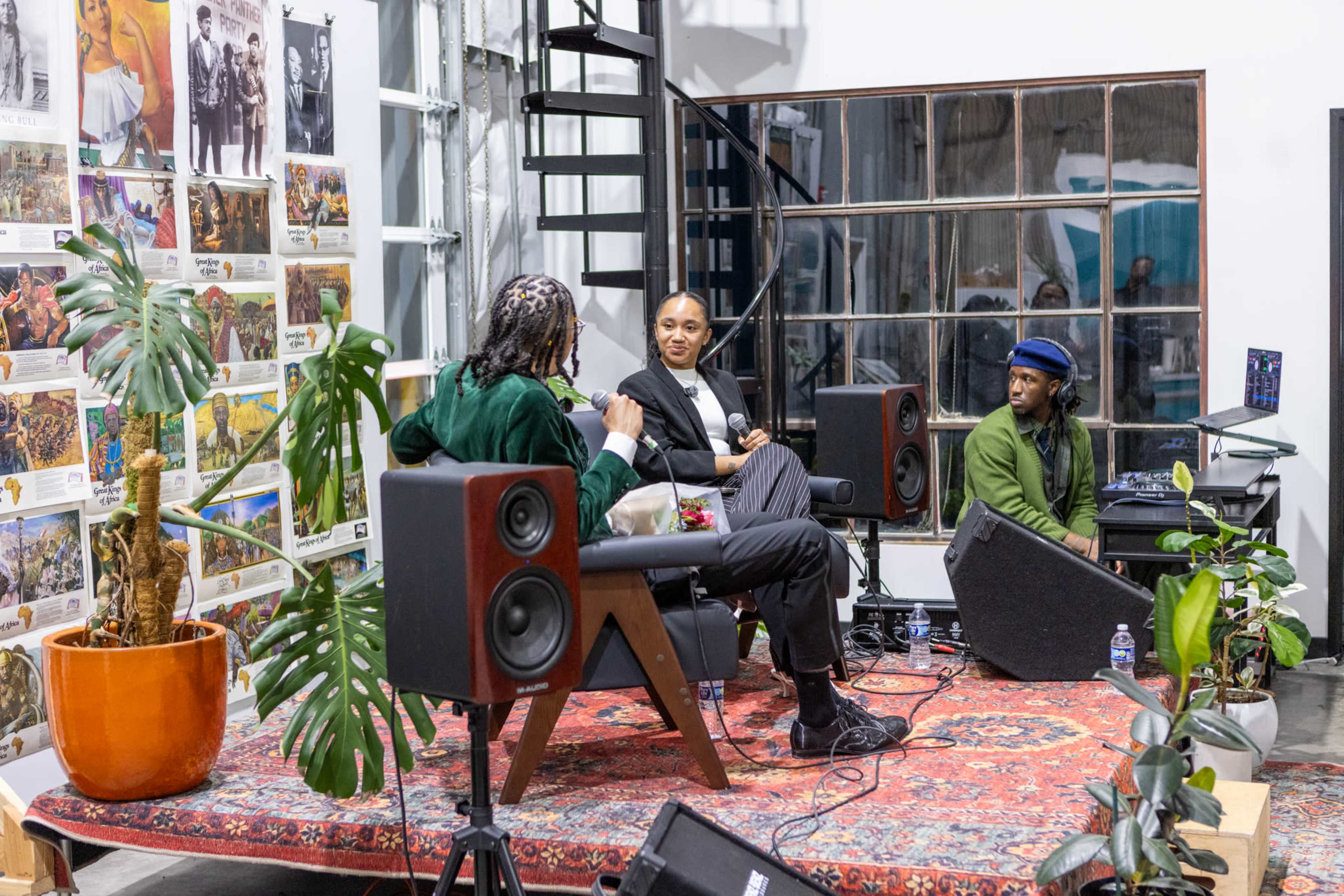 Three individuals are seated in a discussion setting with plants and a colorful rug in a room featuring art on the walls.