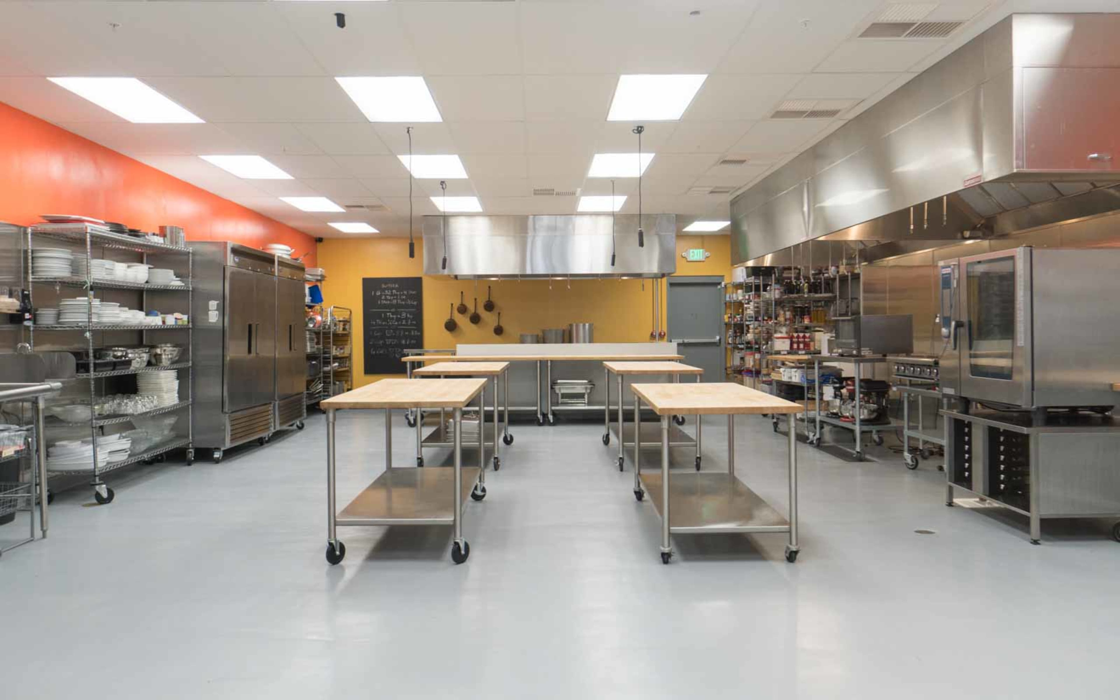 A modern kitchen space with stainless steel appliances, several wooden prep tables, and well-organized storage areas on the walls.