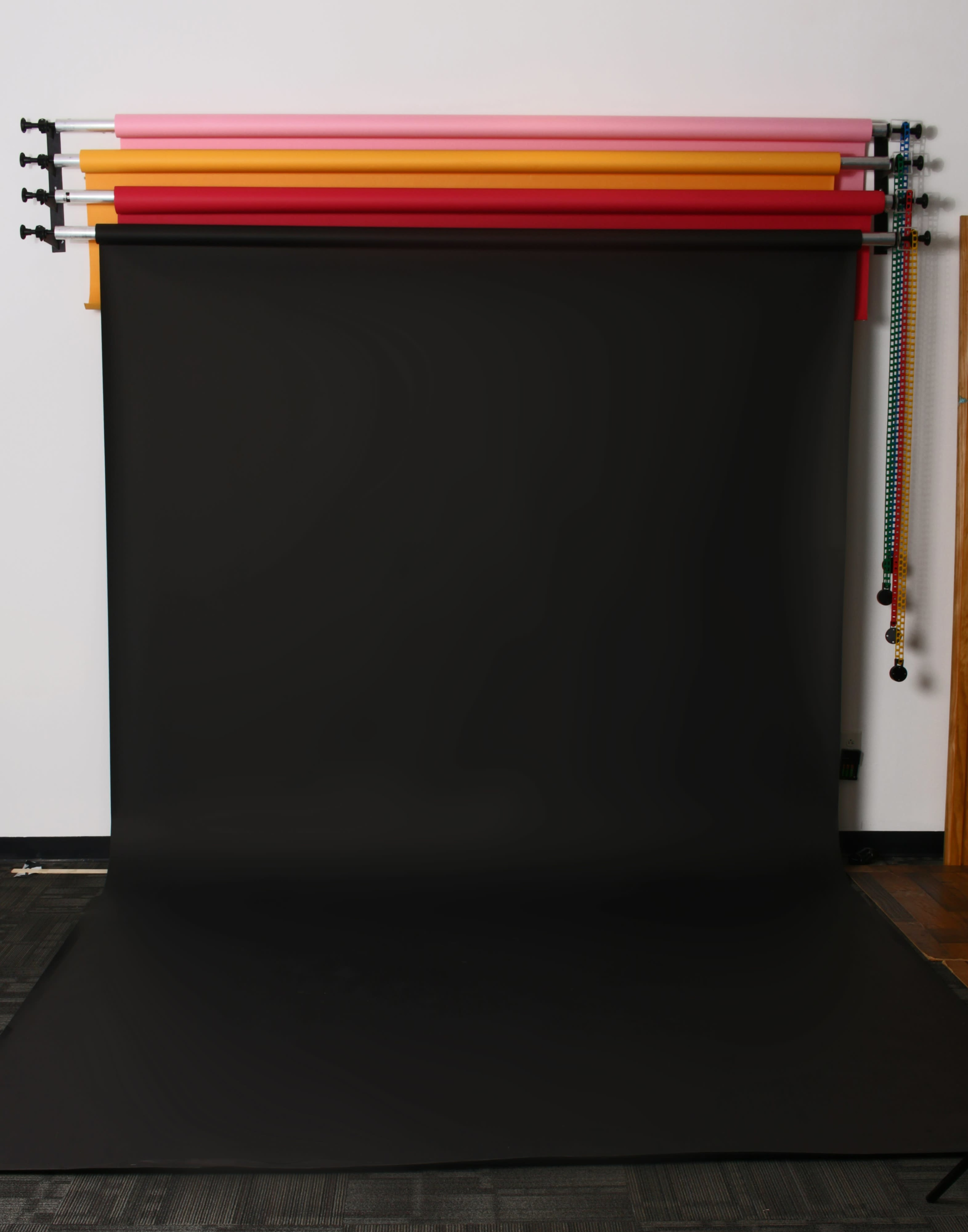 A studio backdrop setup features a black fabric roll with multiple colored paper rolls mounted above it.