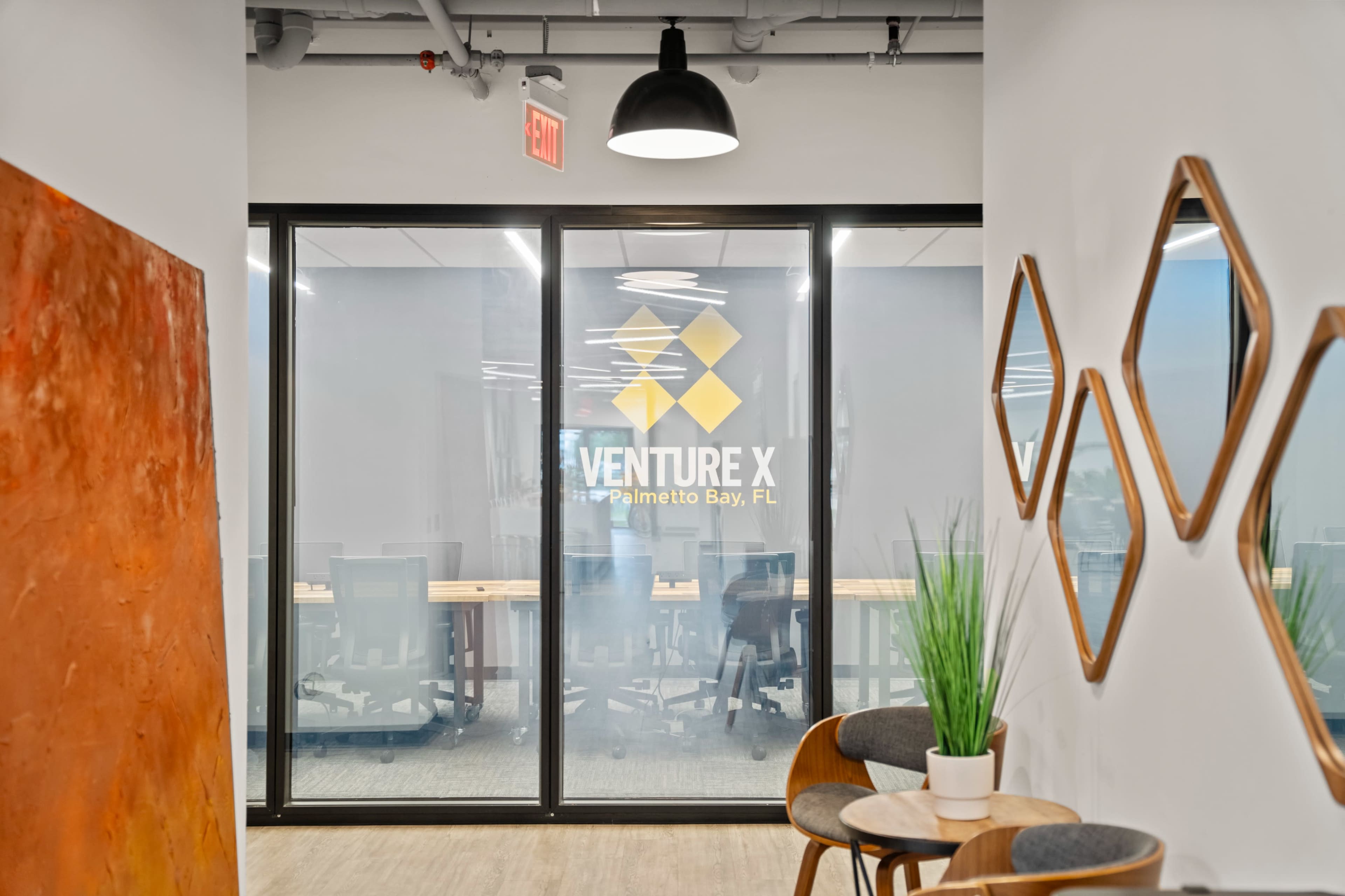 The image shows a modern office corridor with glass walls displaying the "Venture X" logo, accompanied by decorative plants and wall decor on either side.