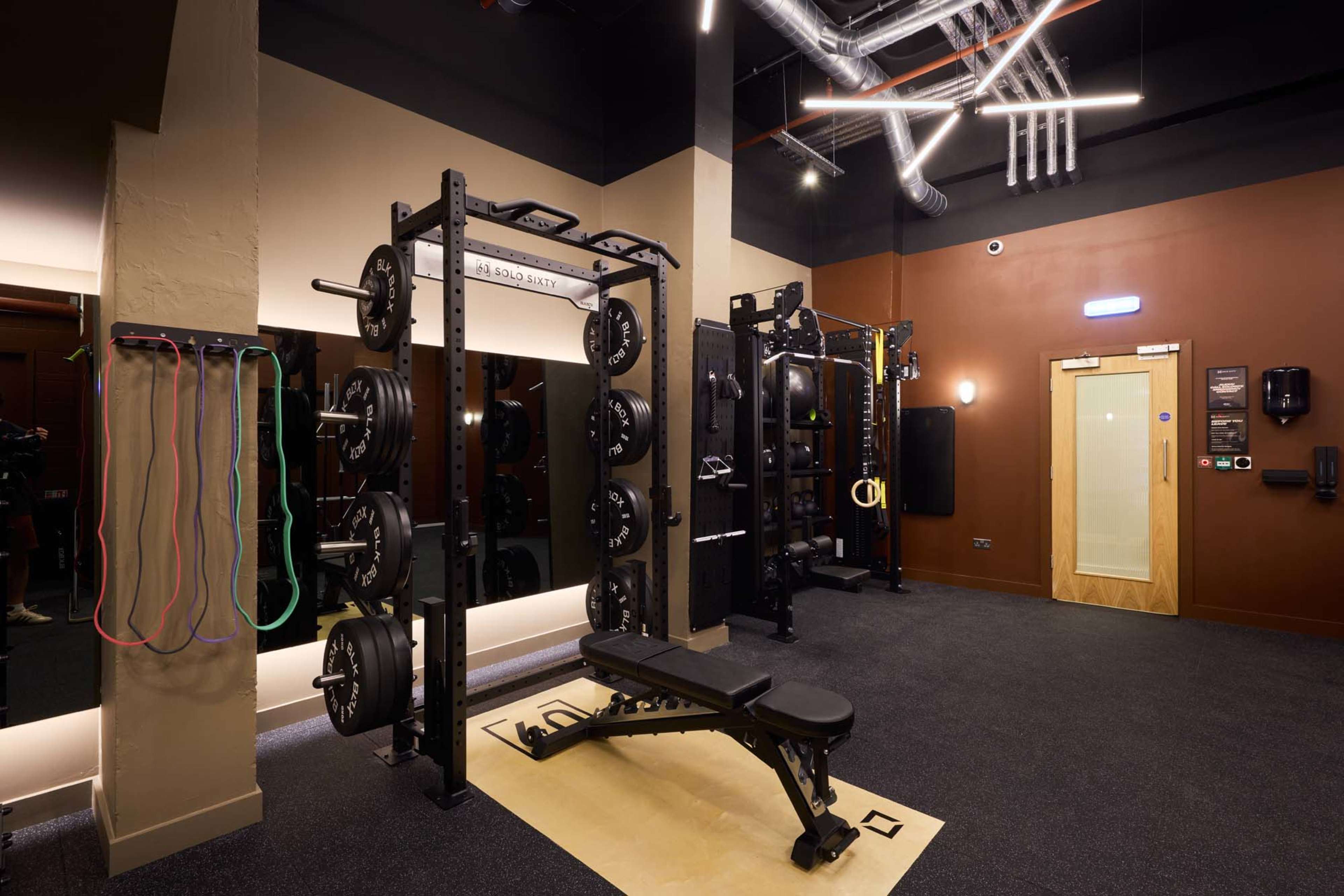 The image shows a modern gym interior with weightlifting equipment, a bench press, and resistance bands hanging on the wall.
