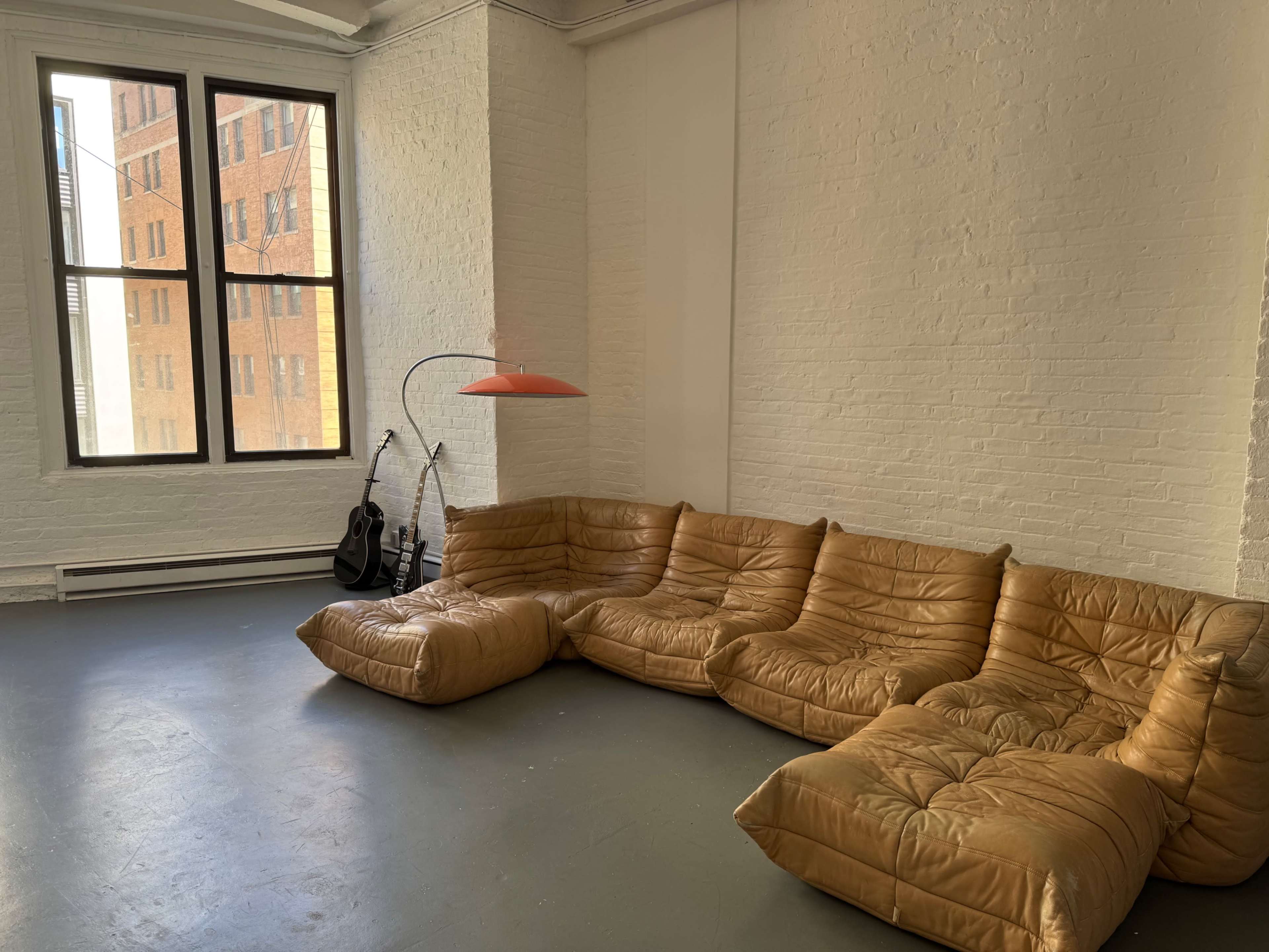The image shows a spacious room featuring a large, tan leather sectional couch, a standing lamp with an orange shade, and two guitars resting against the wall near a large window.