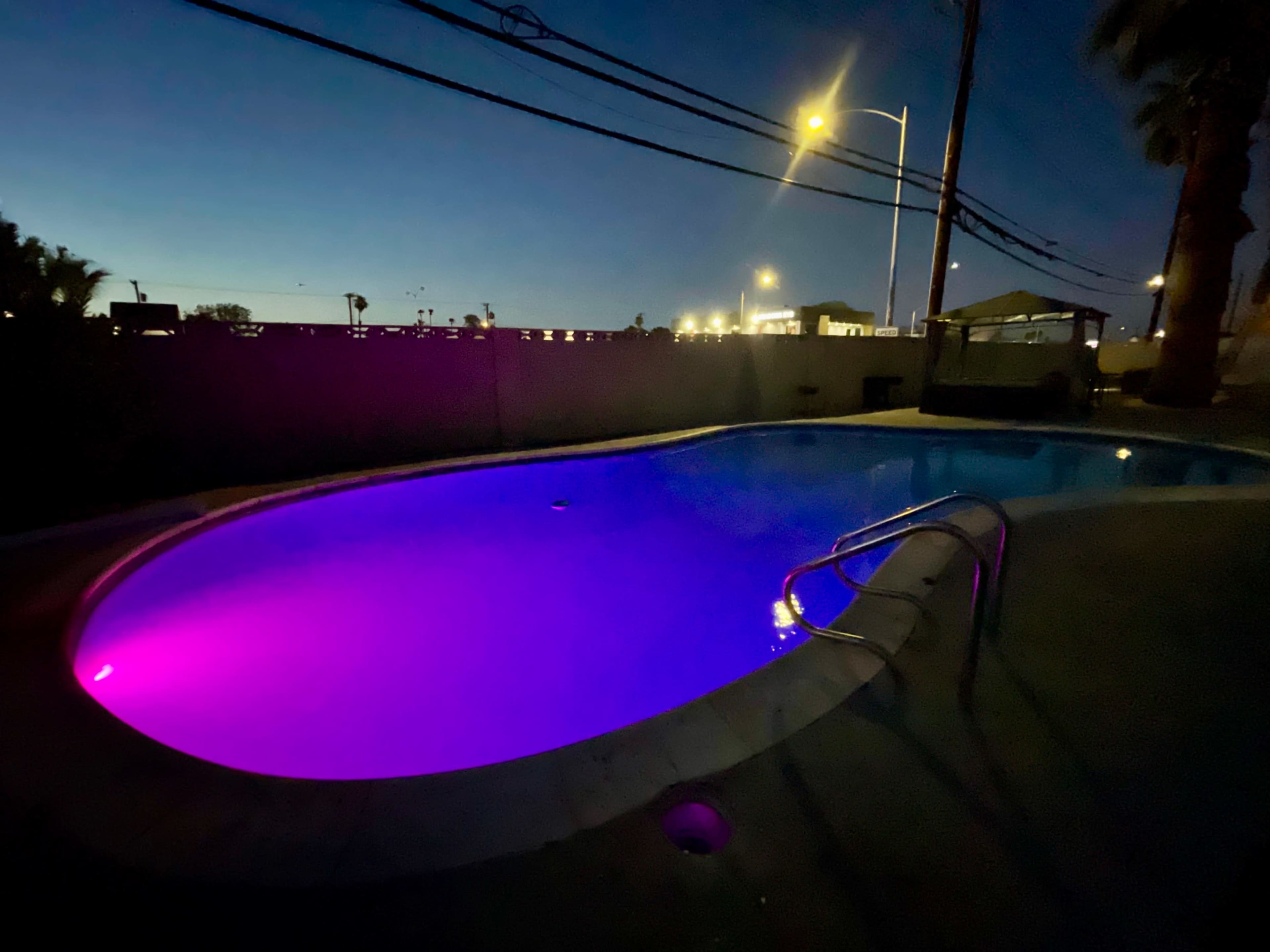A swimming pool illuminated with purple lights sits under a night sky, surrounded by palm trees and distant city lights.