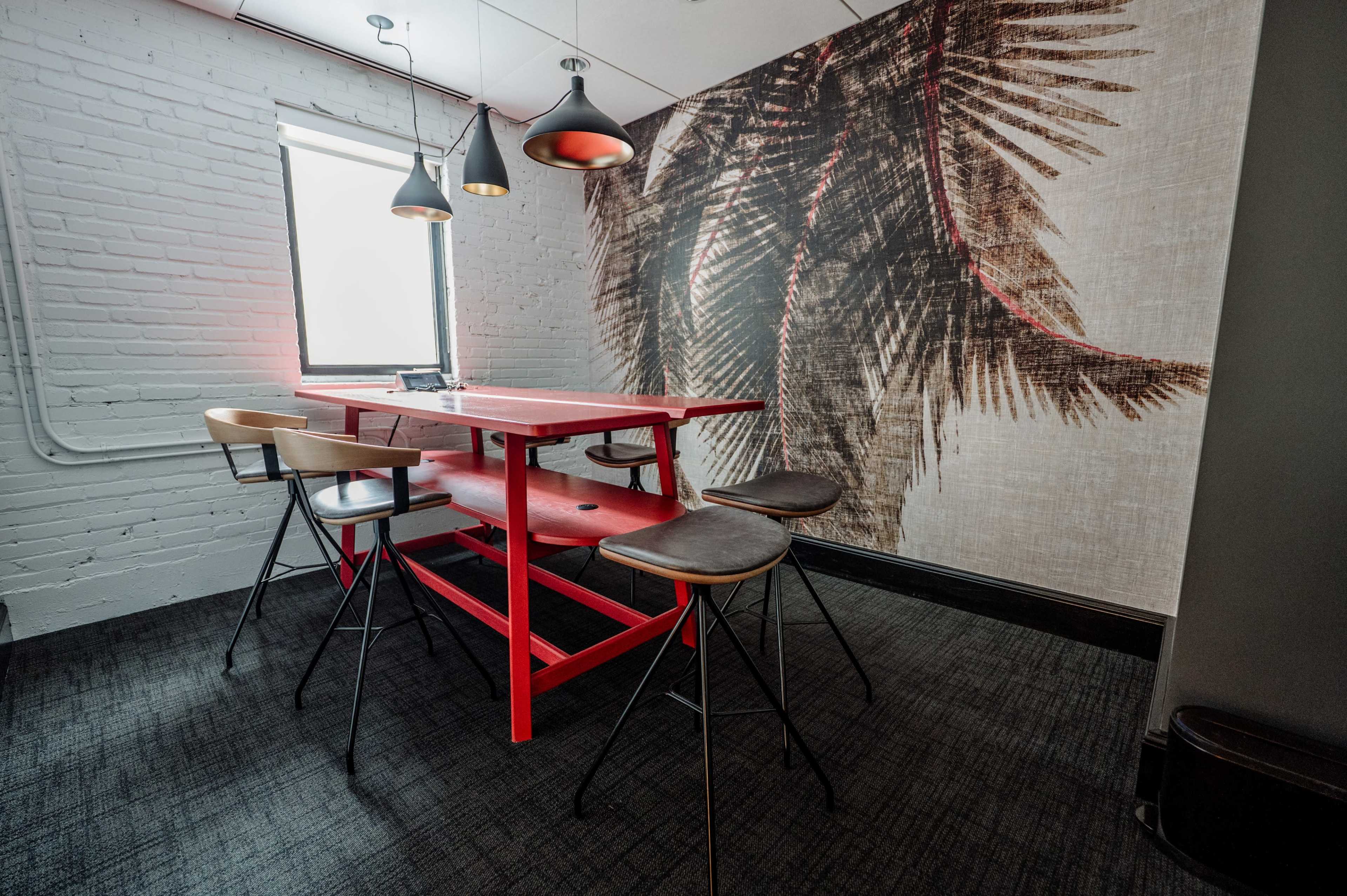The image features a modern workspace with a red table and stools against a wall decorated with a large, textured mural.