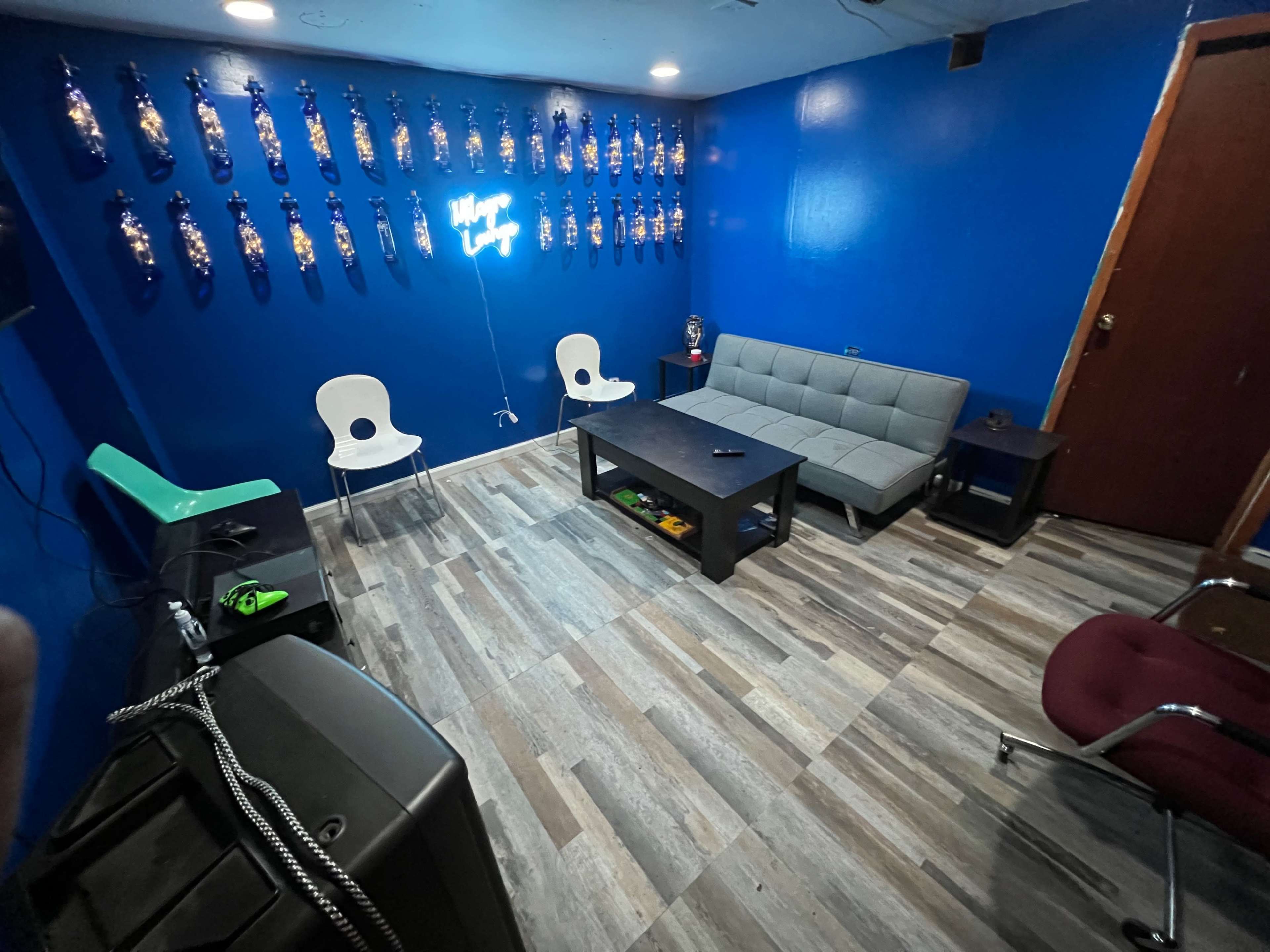 The room features a blue wall adorned with decorative bottles, a gray sofa, two white chairs, and a dark coffee table on a wood-patterned floor.