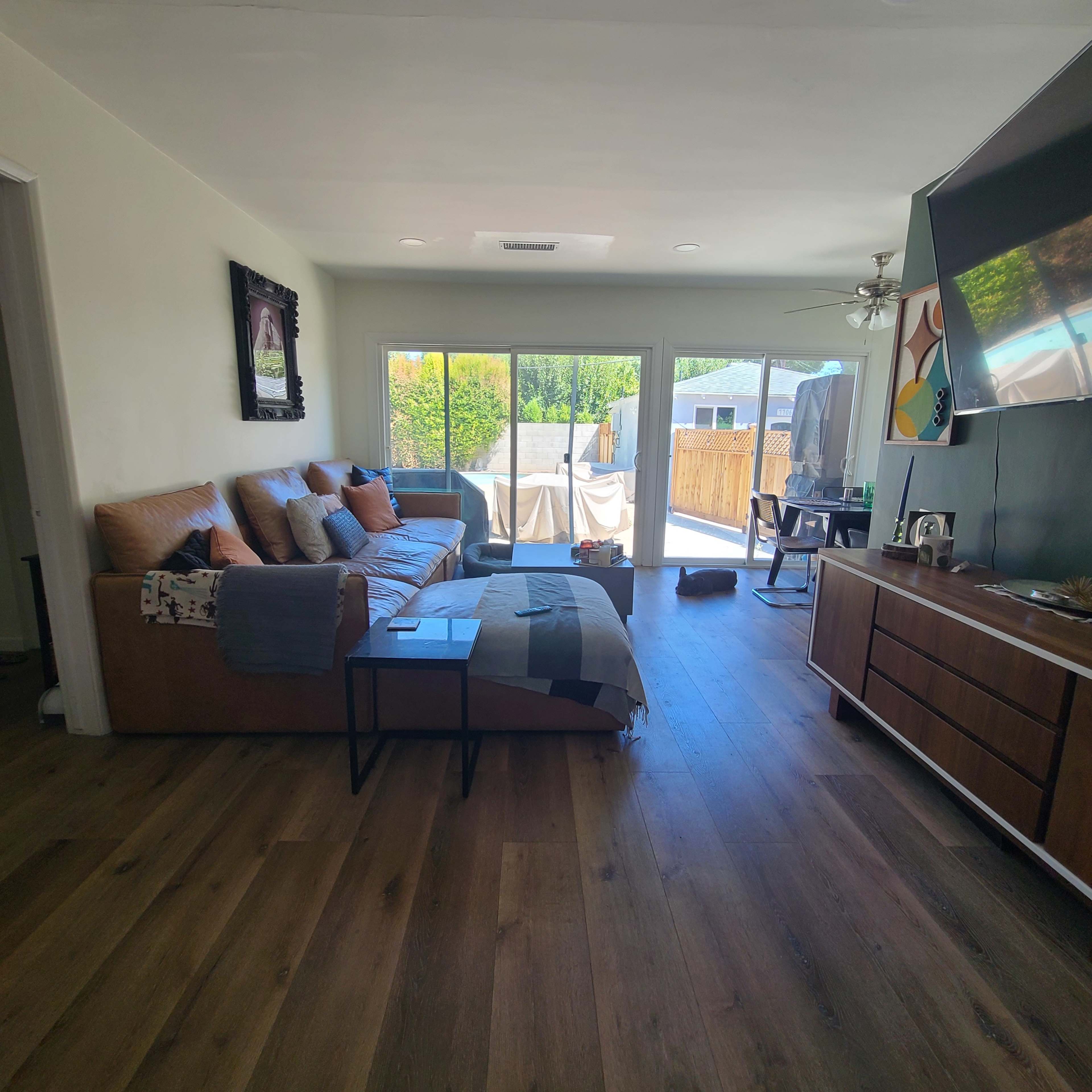 The image shows a living room with a brown leather sofa, wooden flooring, and large sliding glass doors leading to an outdoor area.