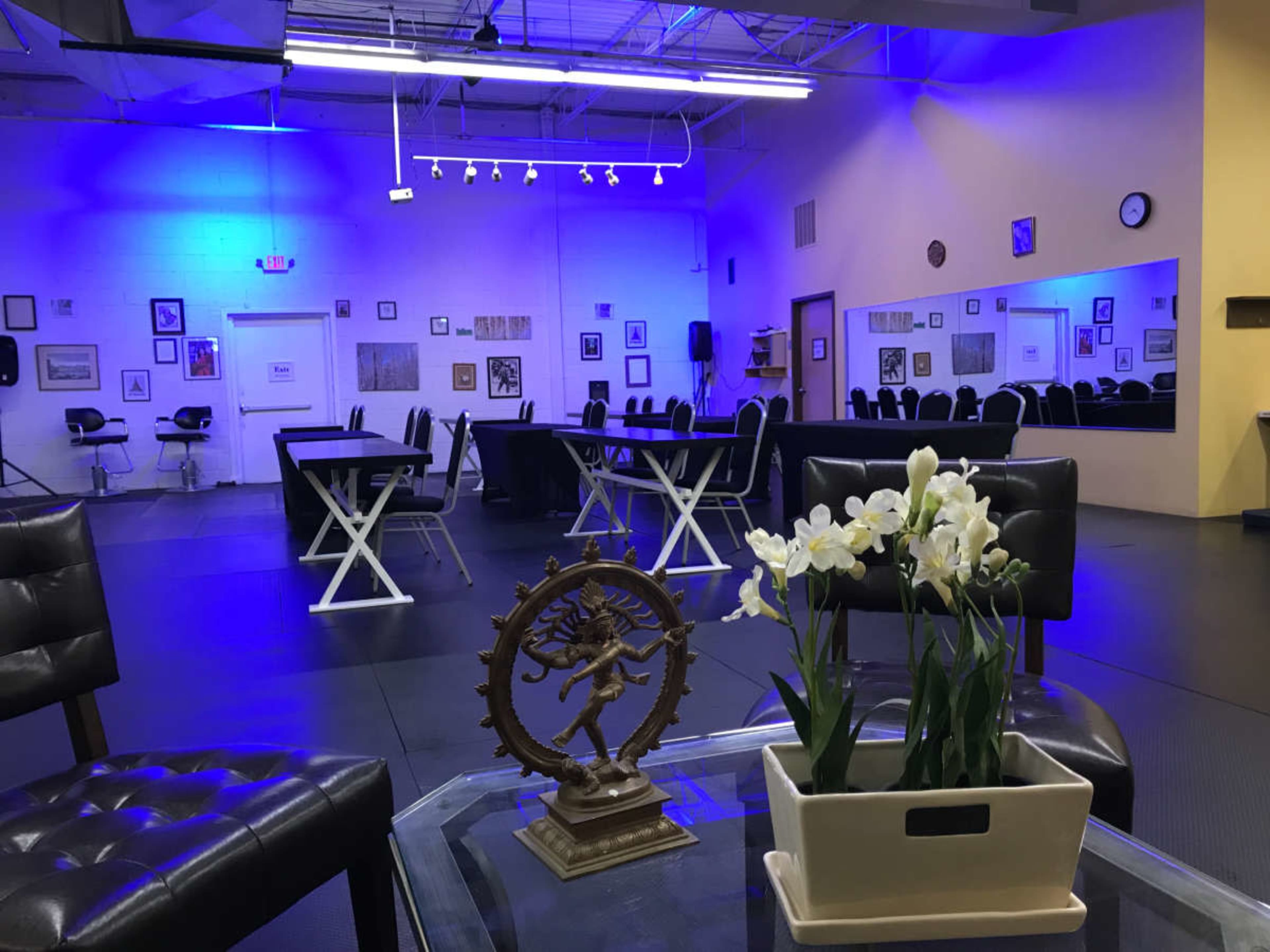 The image shows a spacious room with tables and chairs arranged for an event, illuminated by blue lighting, with a decorative table in the foreground featuring a plant and a sculpture.
