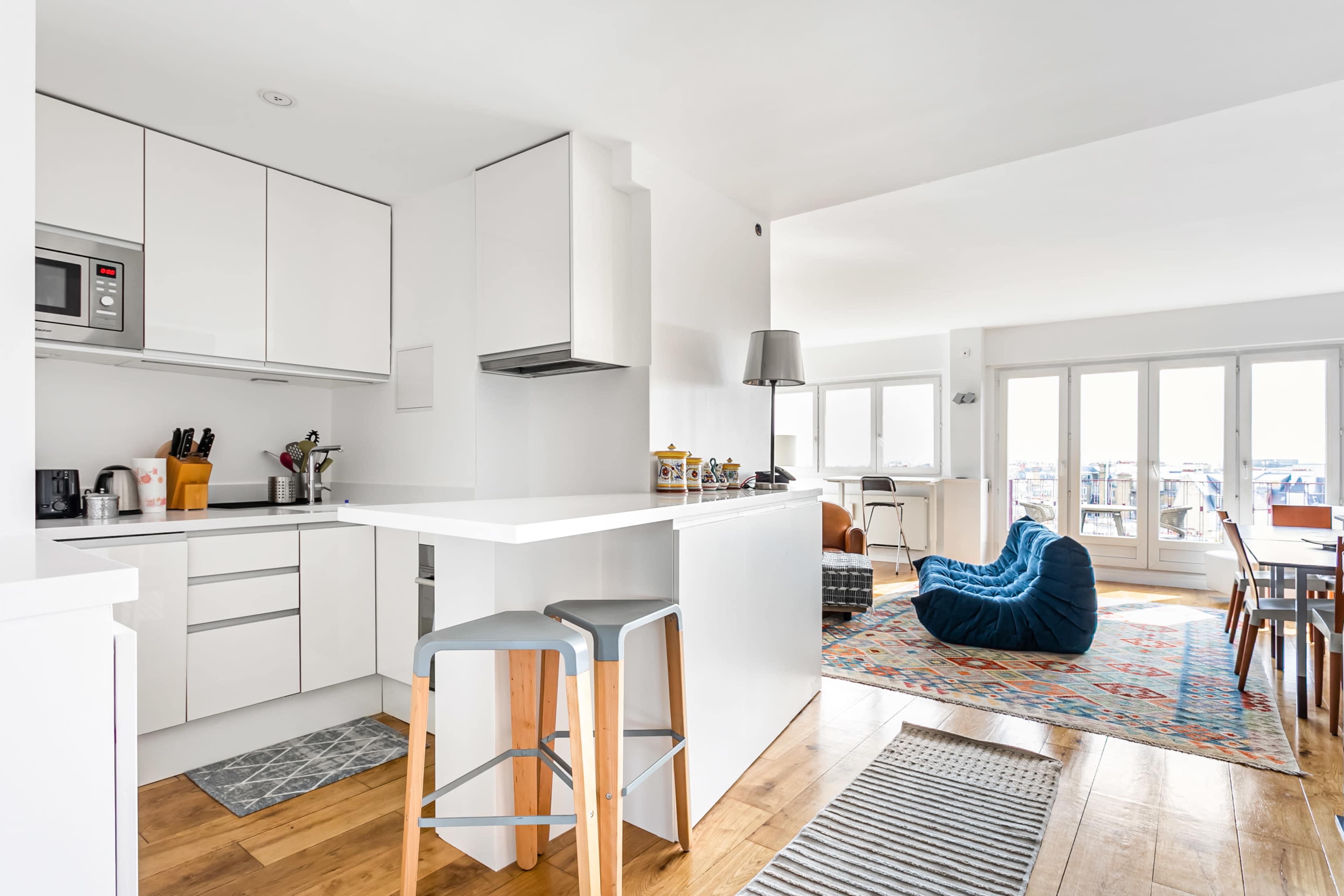 A modern kitchen features white cabinetry and appliances, with a bar area and stools, overlooking a spacious living room with a colorful rug and a large window.