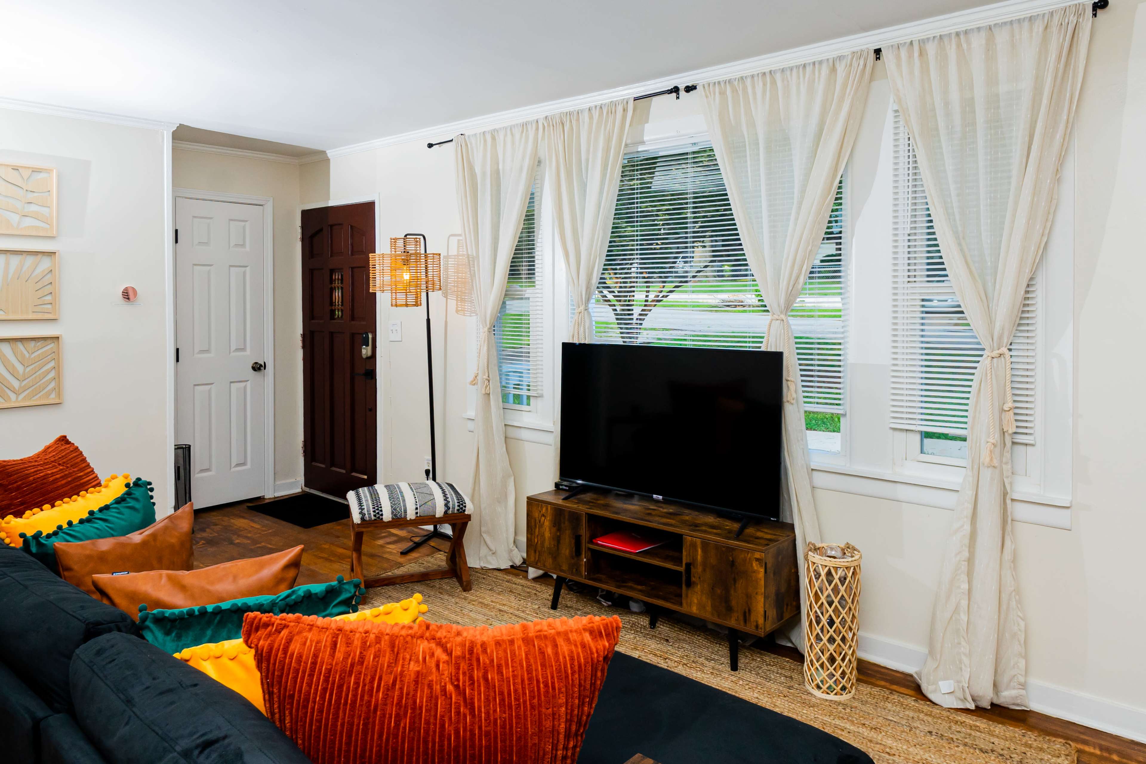 A cozy living room features a black sofa adorned with colorful cushions, a wooden TV stand, and large windows dressed with sheer curtains.