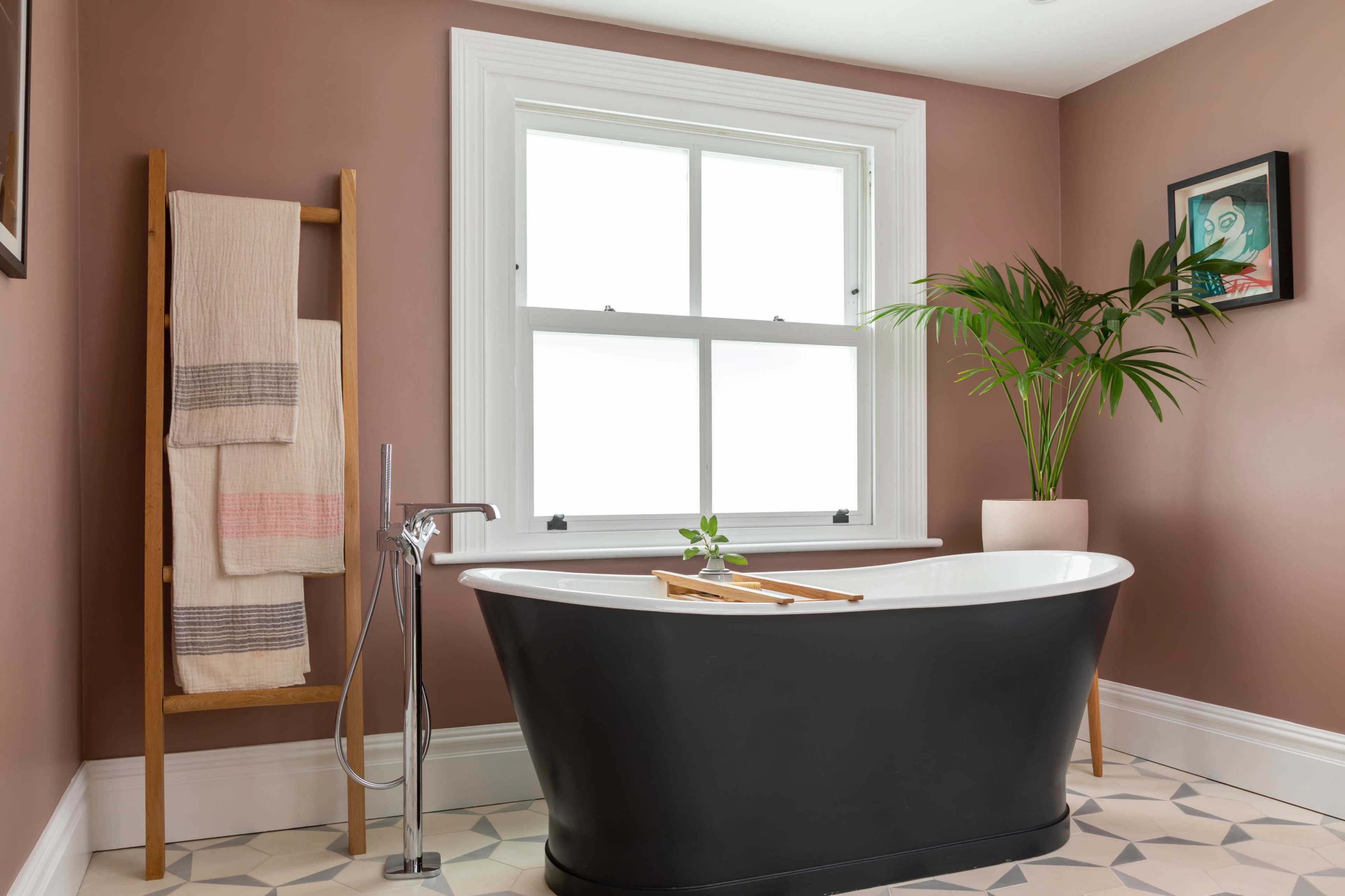 A black freestanding bathtub is positioned beside a large window in a bathroom featuring a beige wall and a plant in a pot.